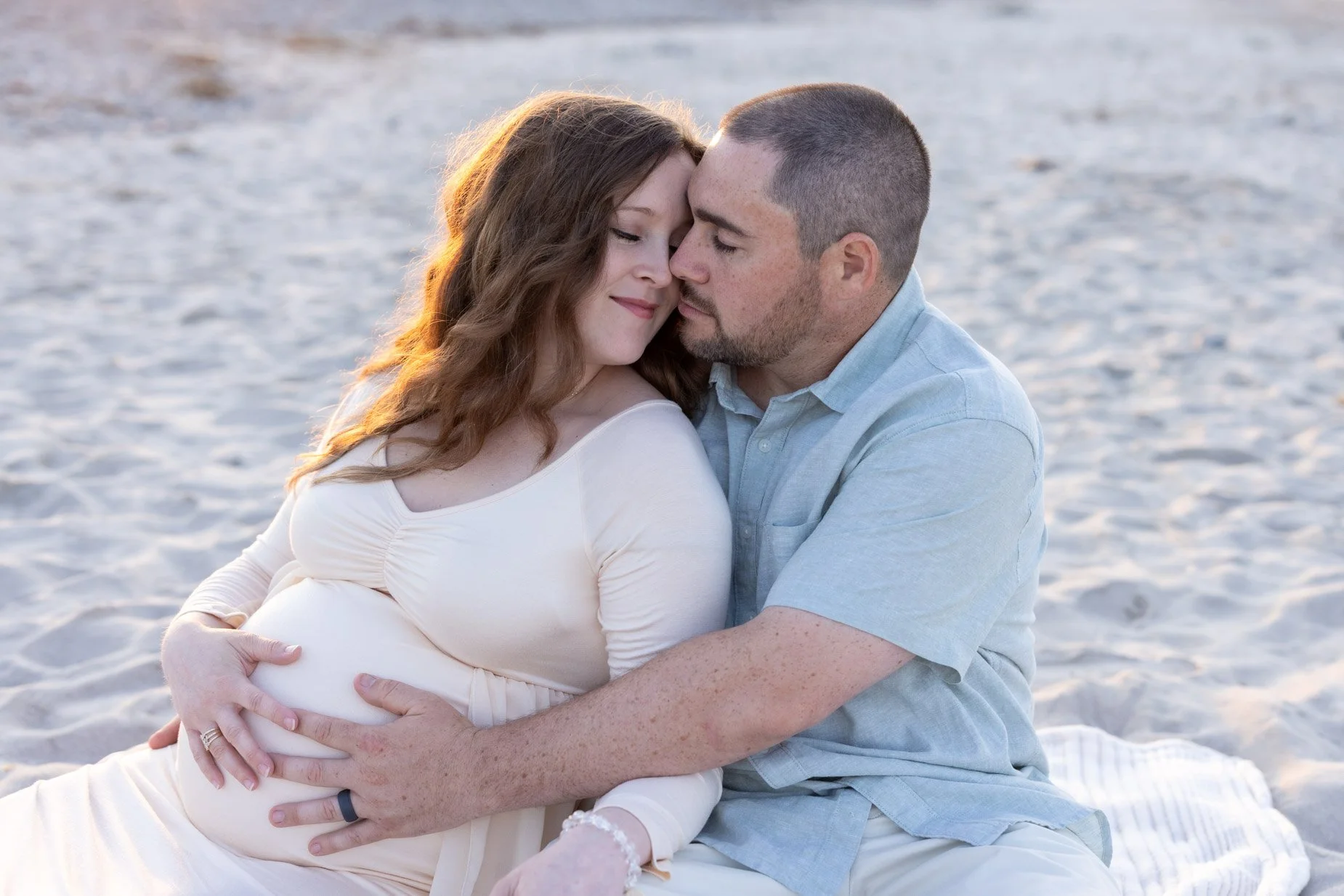 man and expecting woman nuzzle on blanket at beach
