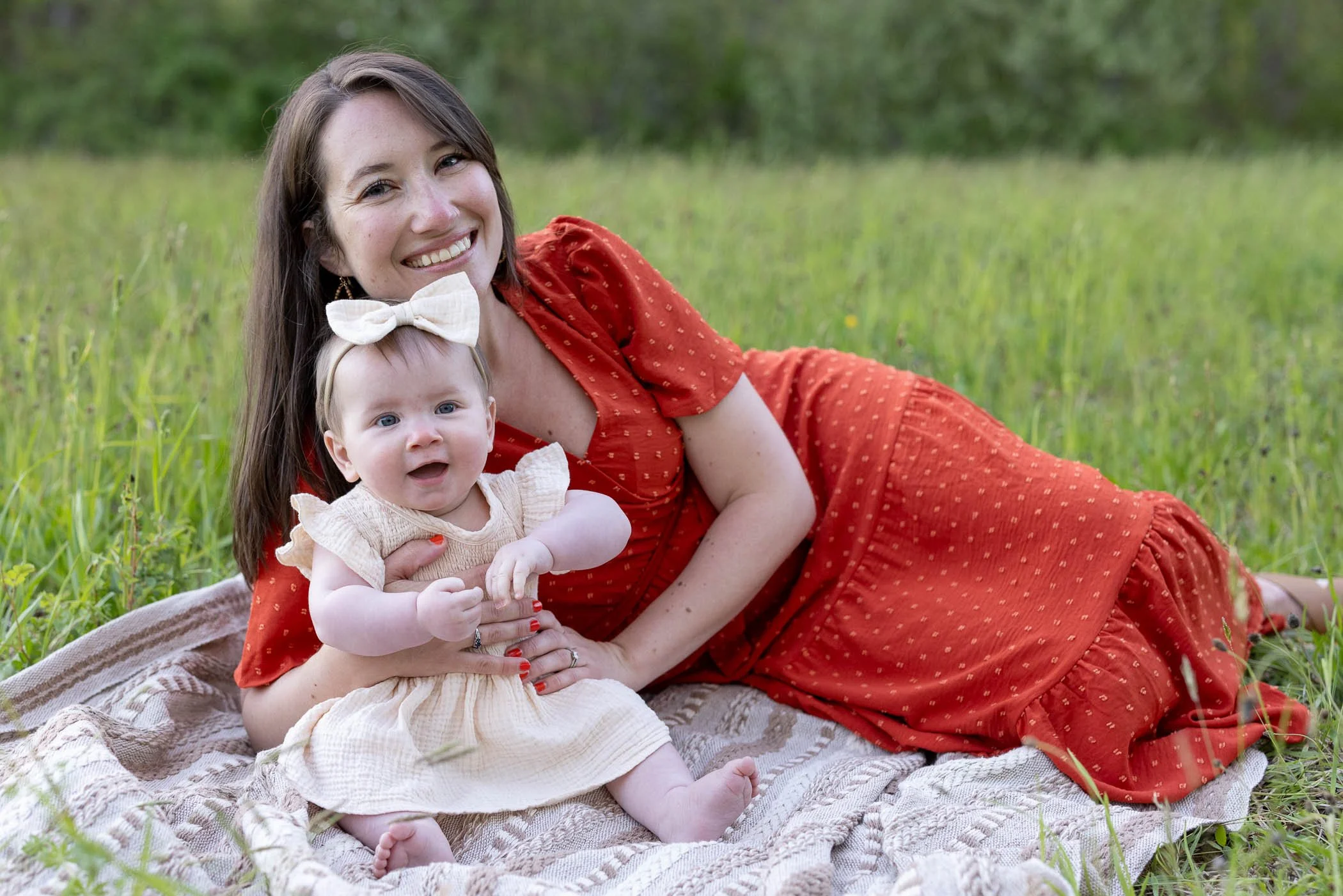 Mom wearing long red dress cuddles sitting baby daugher on blanket in field