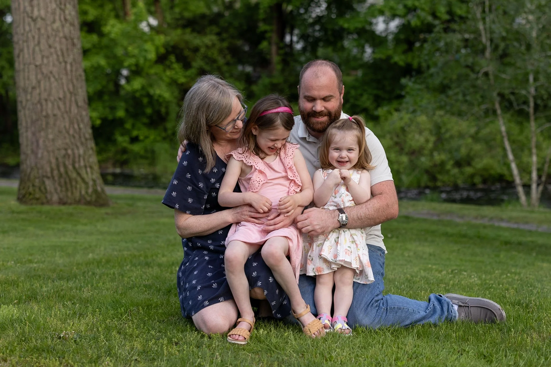 Parents with two small daughters cuddle together in park mom dad tickle little girls everyone laughing showing love and fun