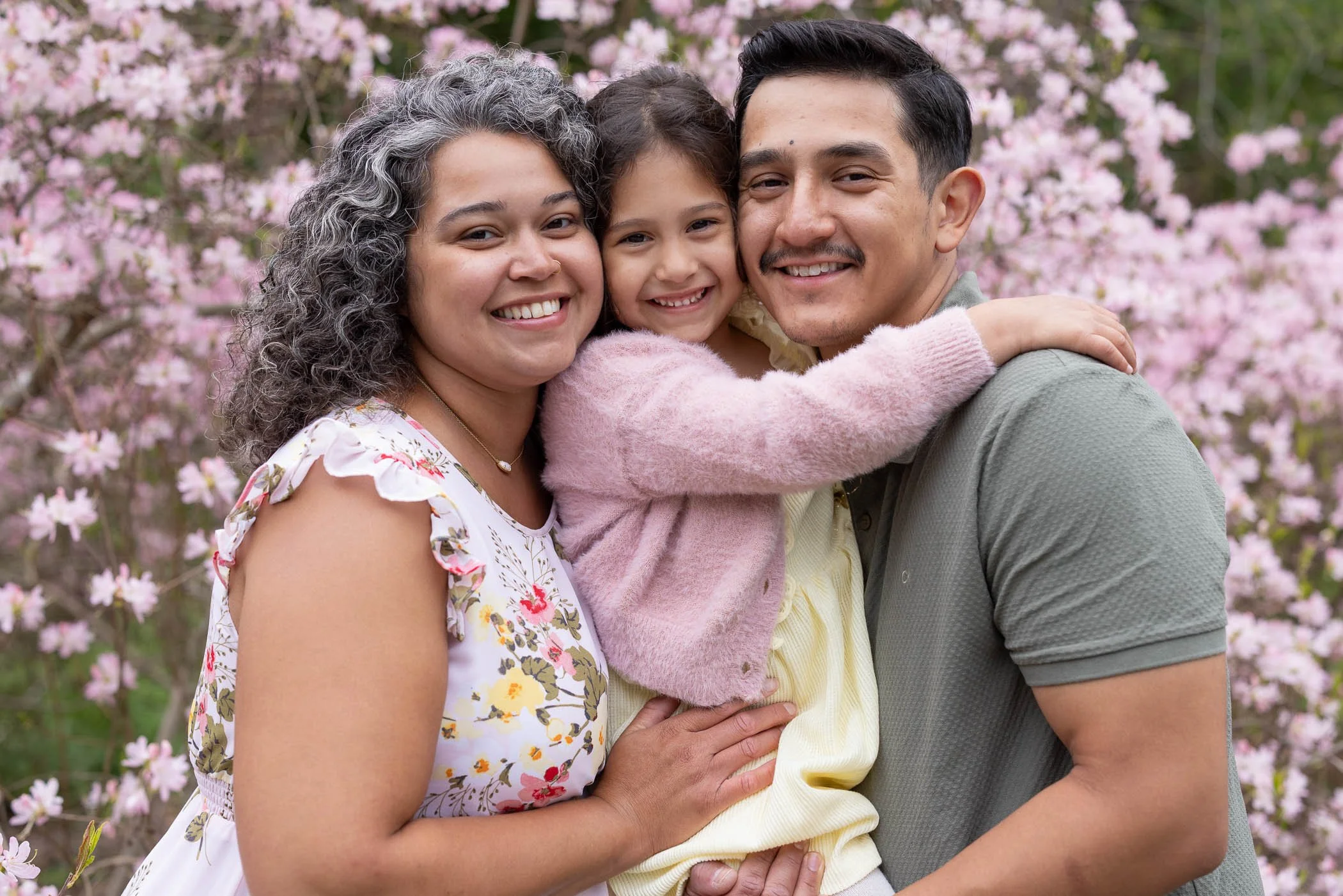 Mom daughter and dad stand cheek to cheek by blooming pink flowers