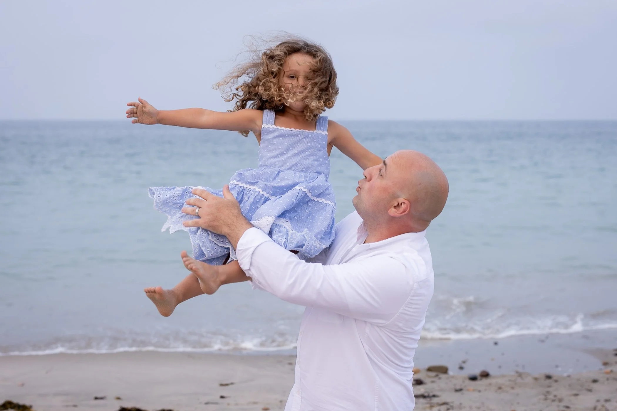 Dad tossing little girl with curly hair wearing blue and white dress in front of ocean showing play and fun