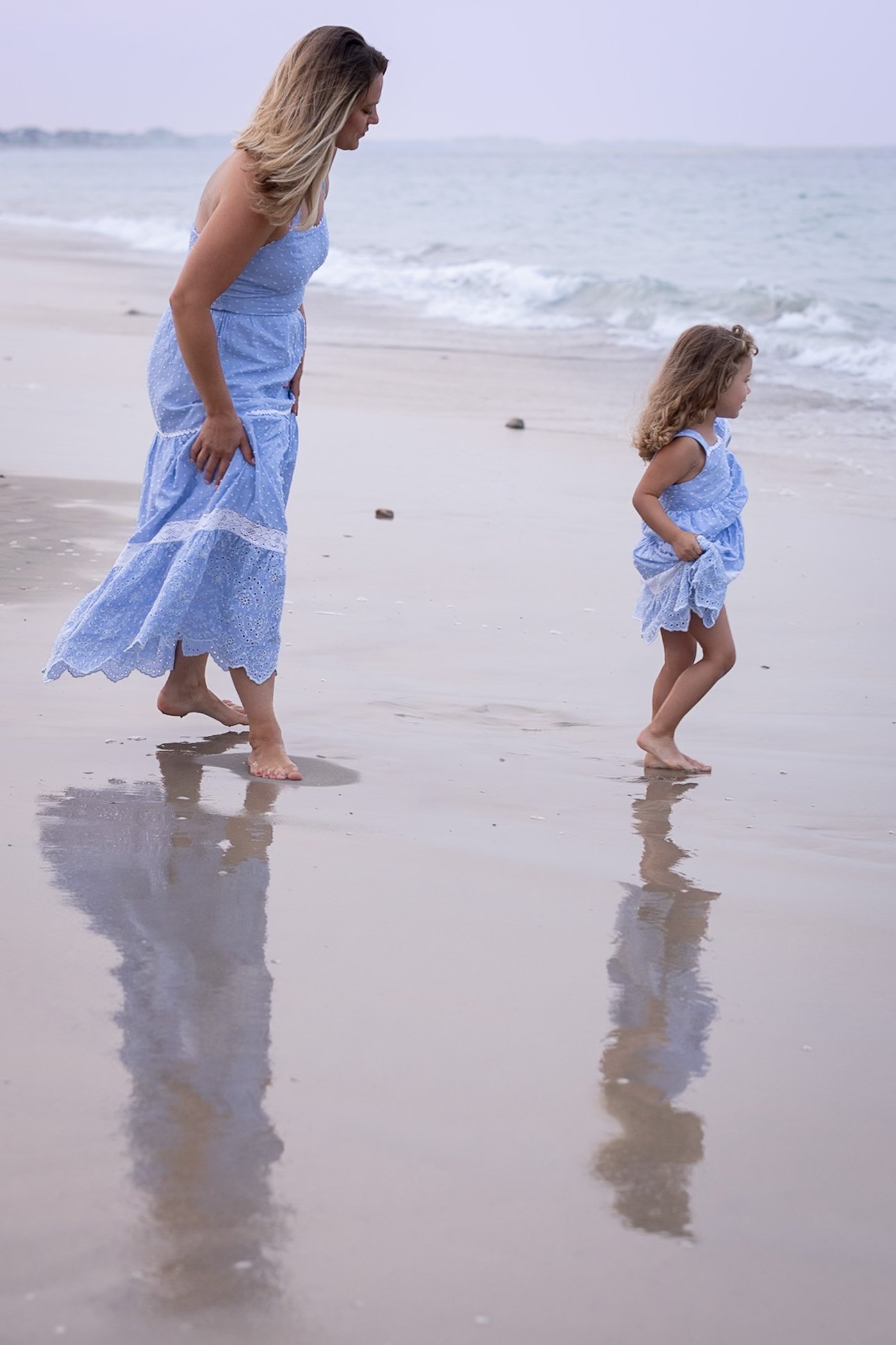 Mom and young daughter wearing pretty blue dresses walk barefoot on the wet sand toward the ocean casting long shadows