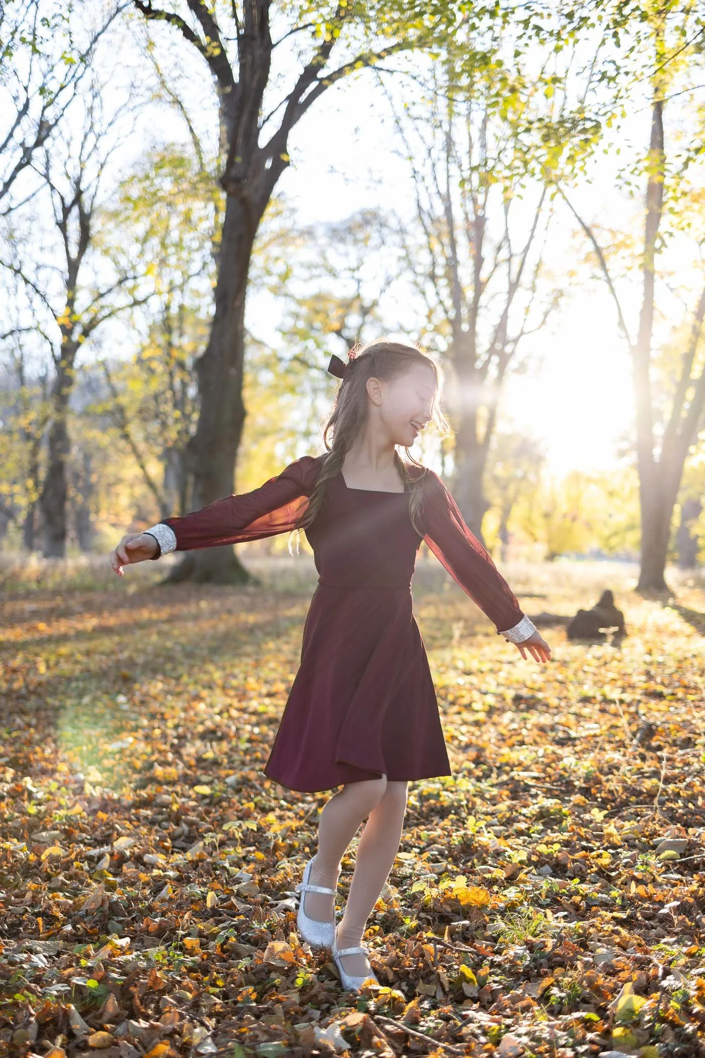Young girl smiling wearing pretty maroon dress  twirling dancing in woods with golden hour backlit sunshine
