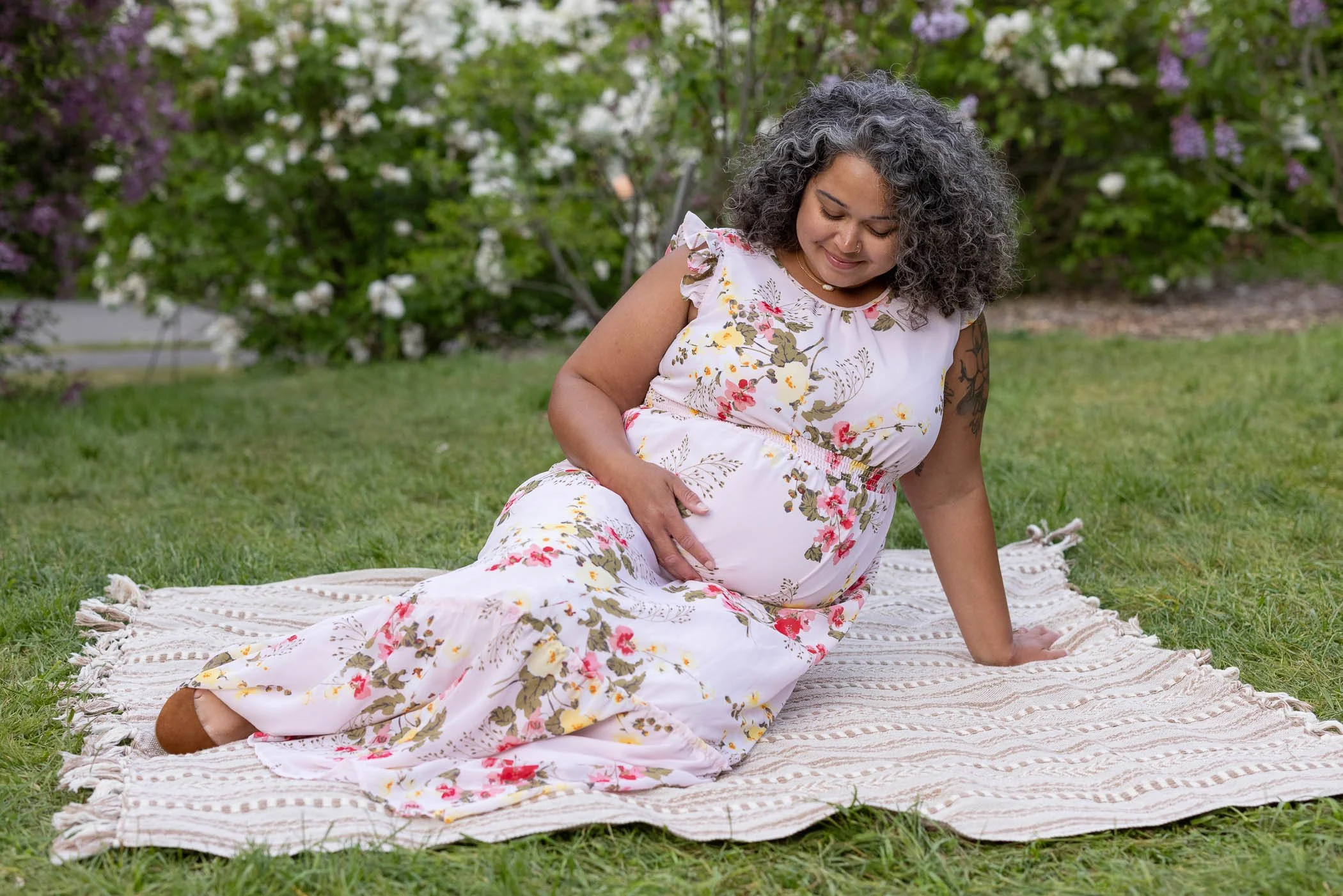 Pregnant lady wearing floral dress sitting on blanket near lilacs
