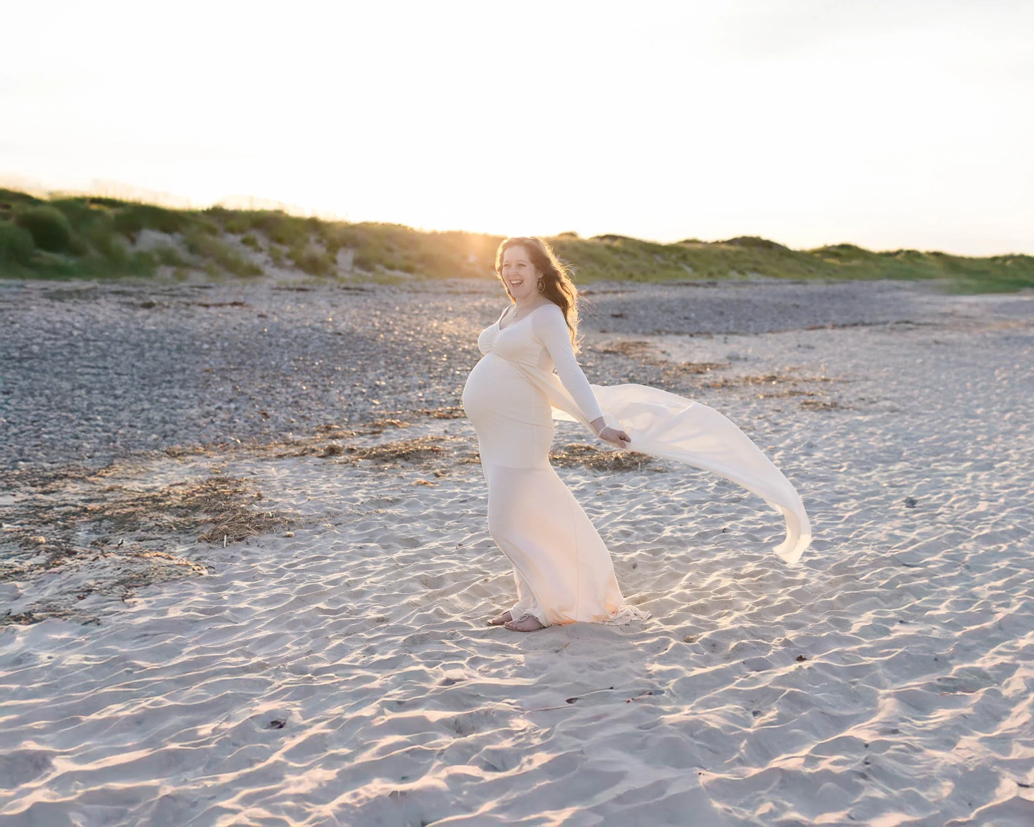 Pregnant woman smiling standing on beach flowing cream color dress
