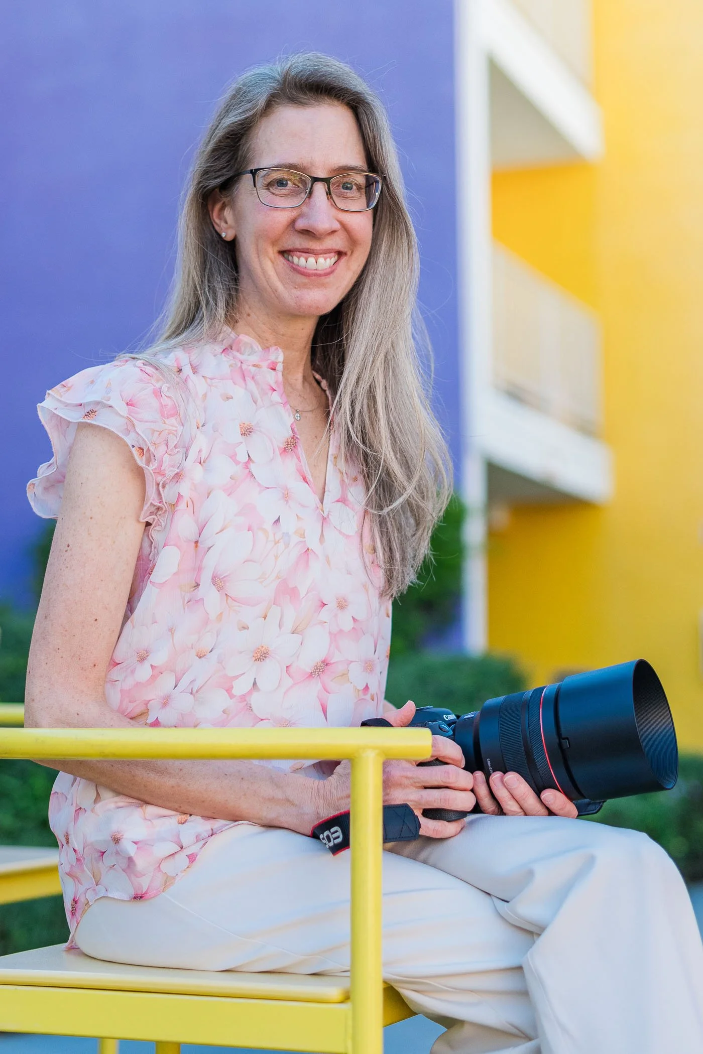 Portrait of Jennifer Banks smiling sitting in yellow chair holding Canon camera wearing pink blouse
