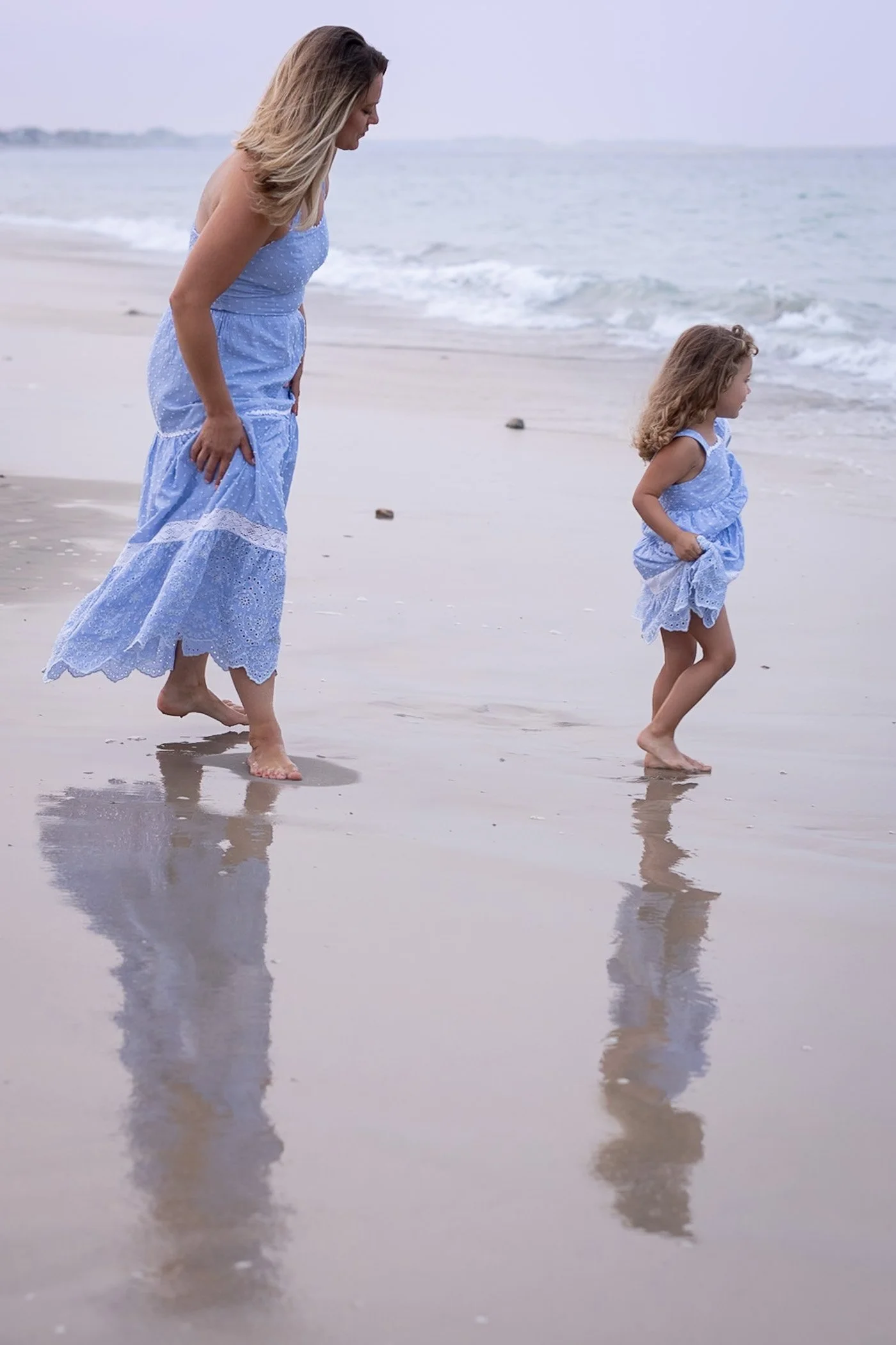 Mom and young daughter wearing pretty blue dresses walk barefoot on the wet sand toward the ocean casting long shadows