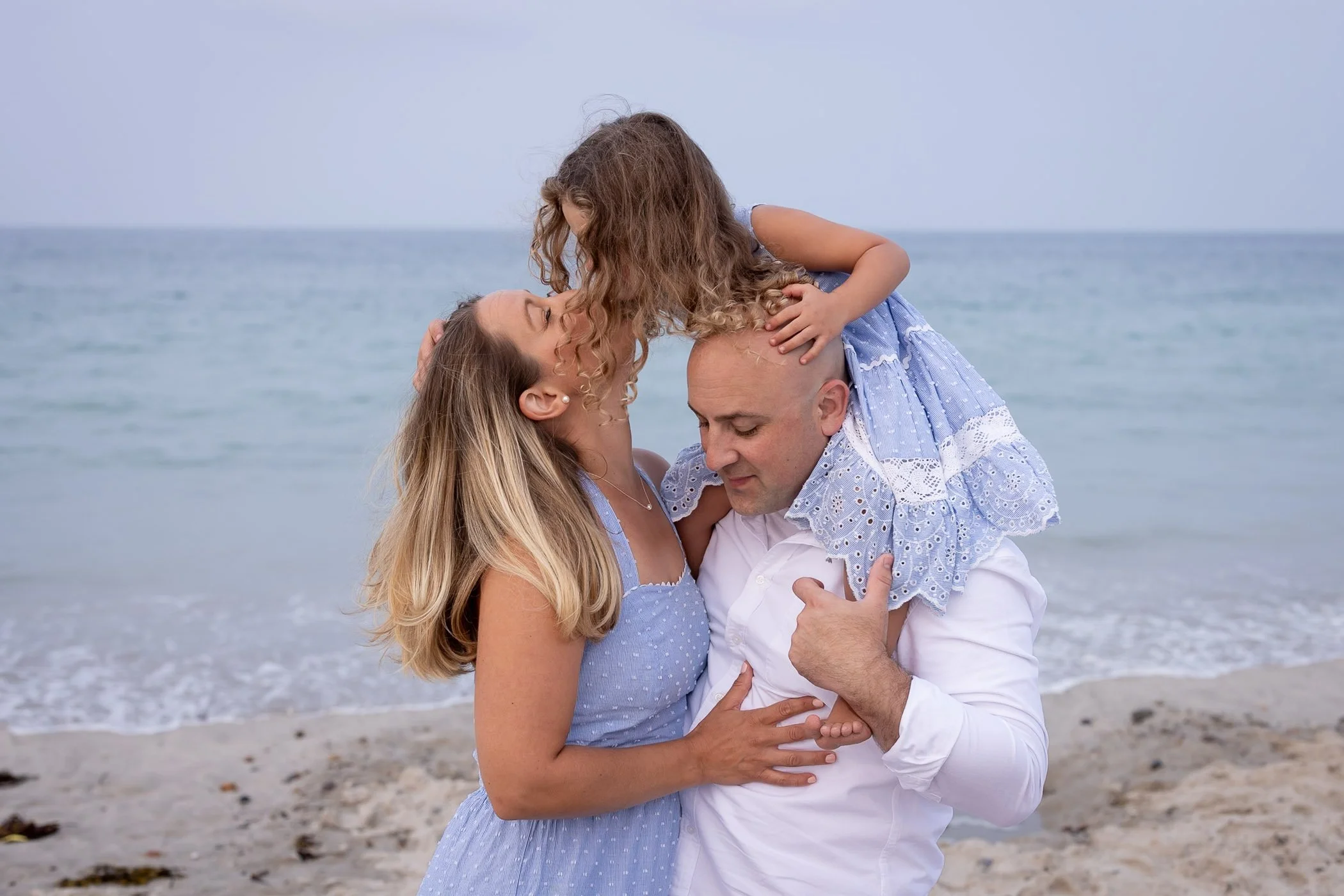 Daughter kissing mom while sitting on dad's shoulders at the beach displaying love and connection
