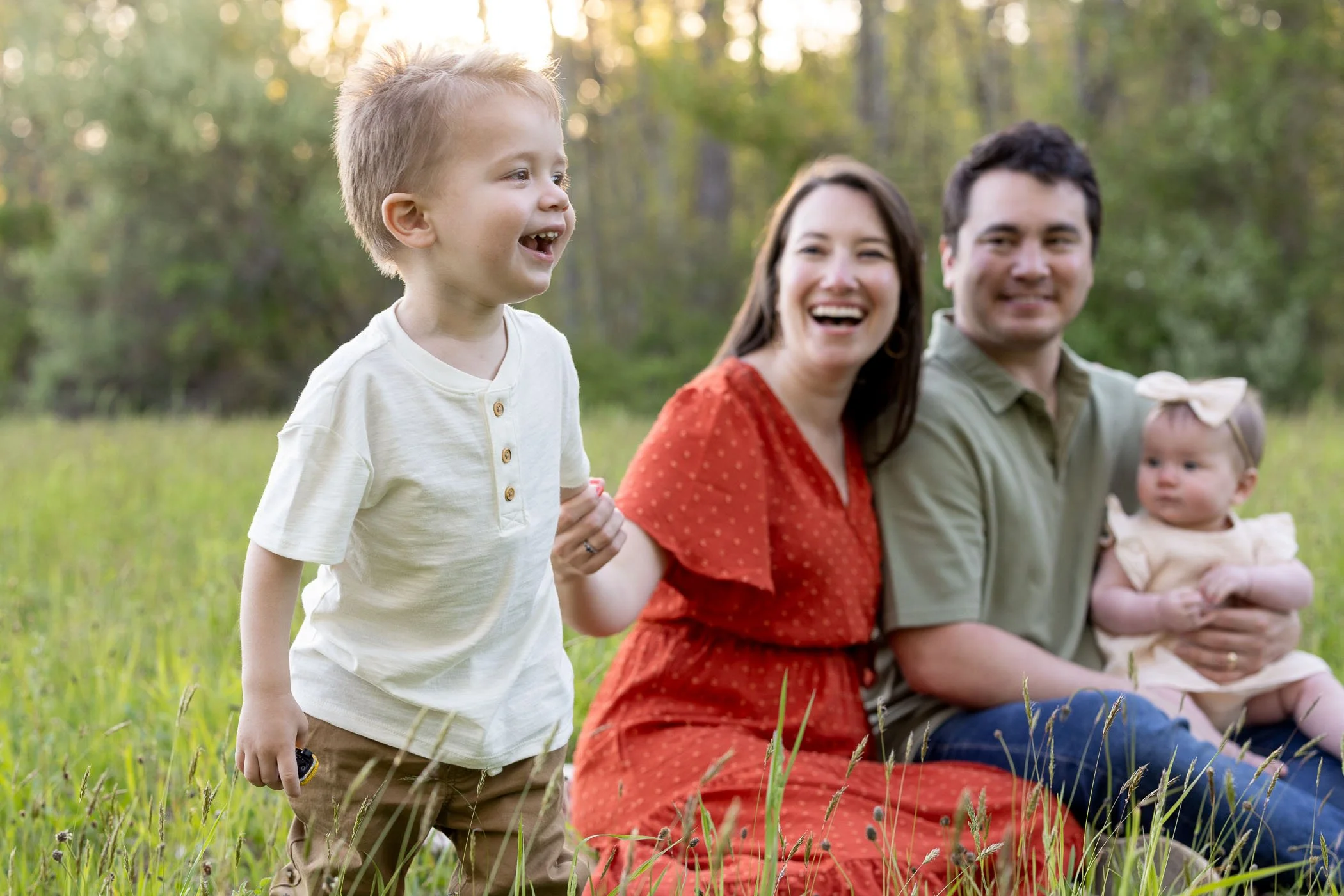 Happy toddler boy stands next to sitting mom, dad, and baby sister in field