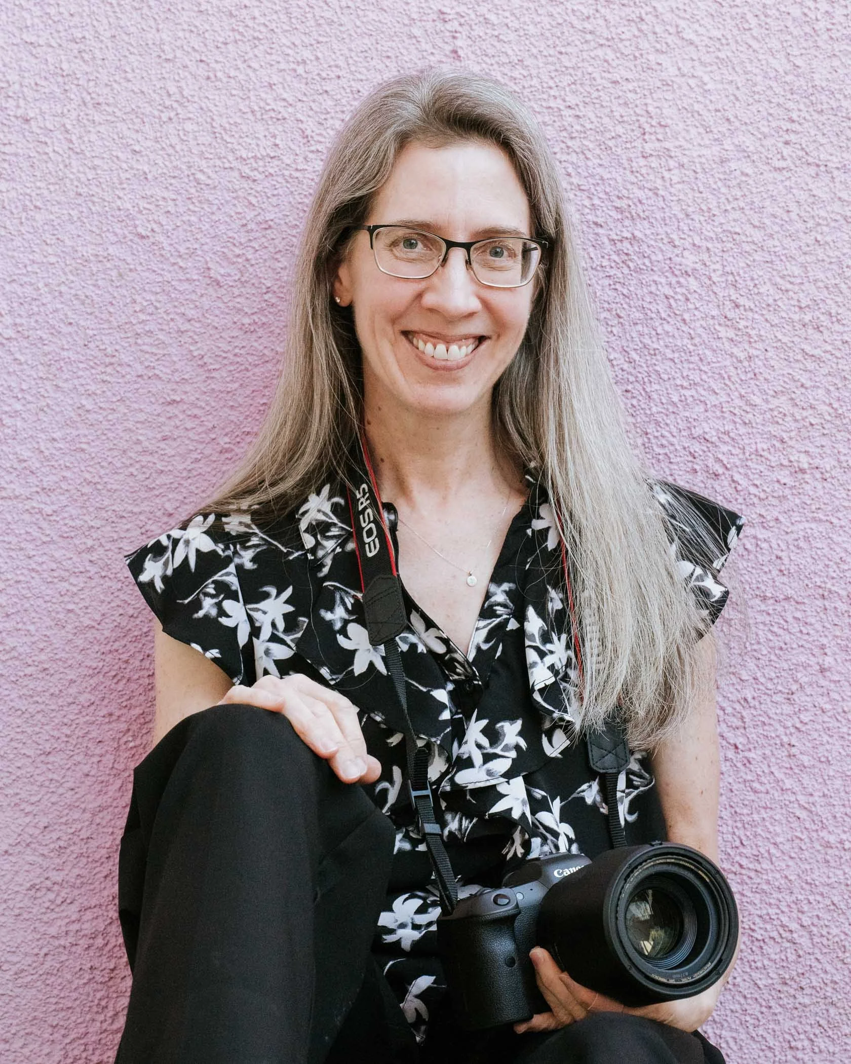 Happy Jennifer Banks sits against lavender wall holding camera