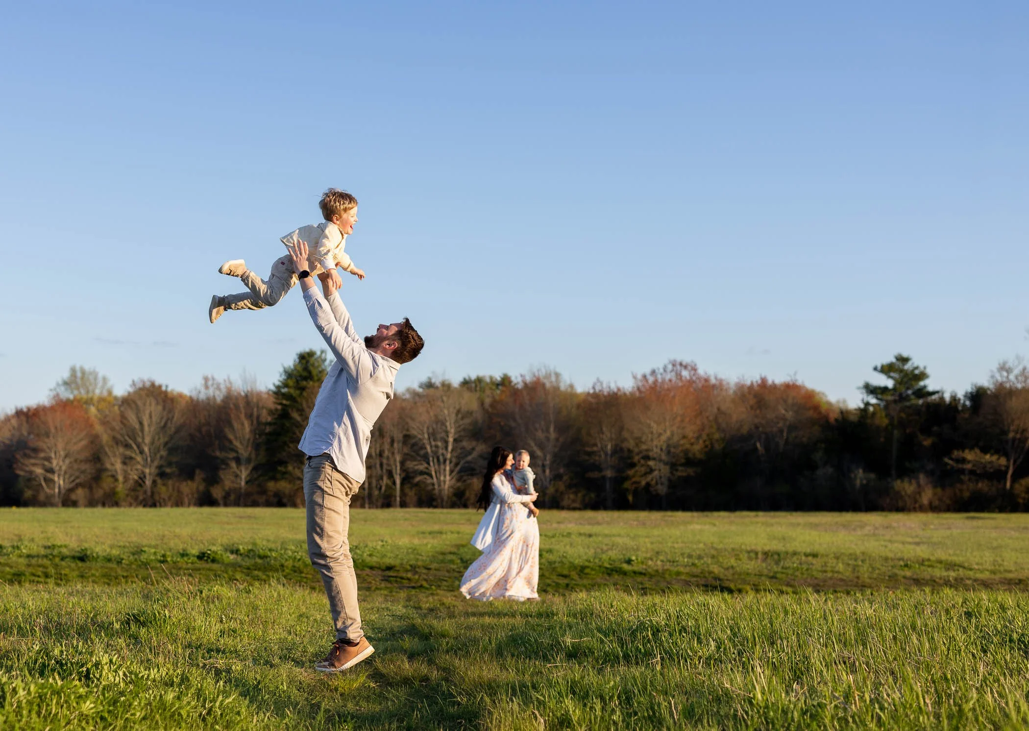 In wide open field, dad tossed little boy into the air while mom cuddles baby