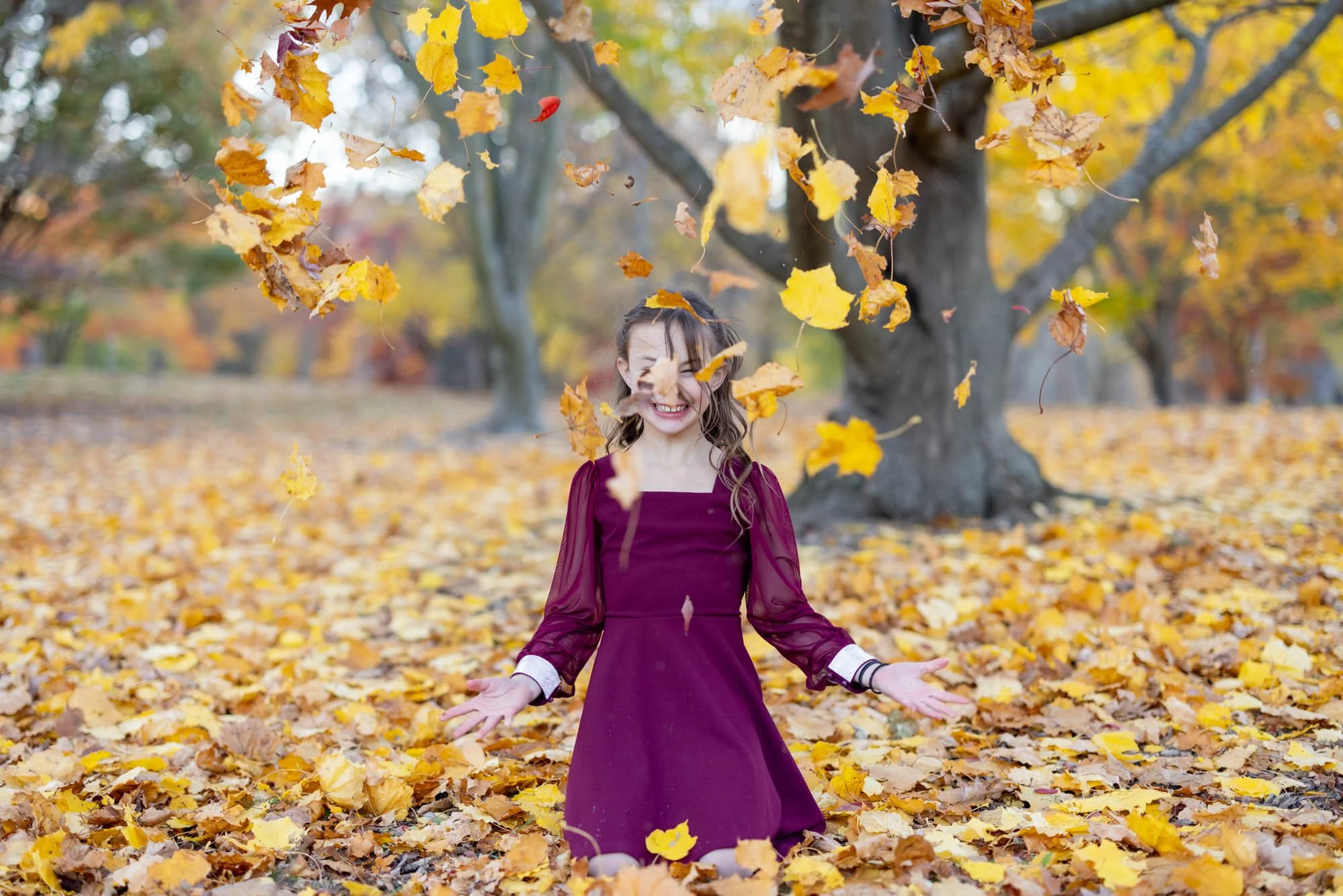girl kneeling down in maroon dress throwing yellow leaves
