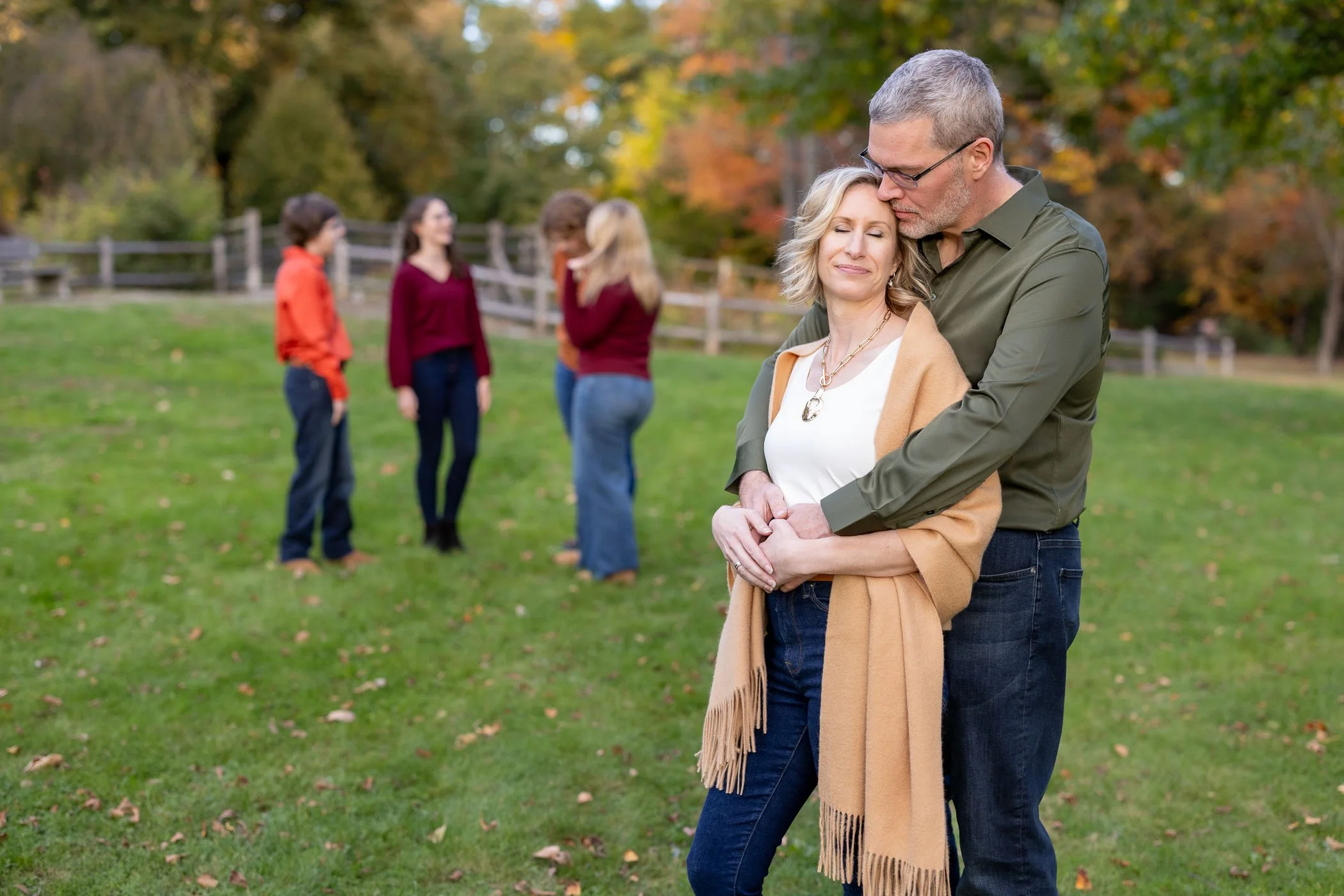 Husband and wife snuggle together with eyes closed in a park while their 4 adult children talk and smile in the background.