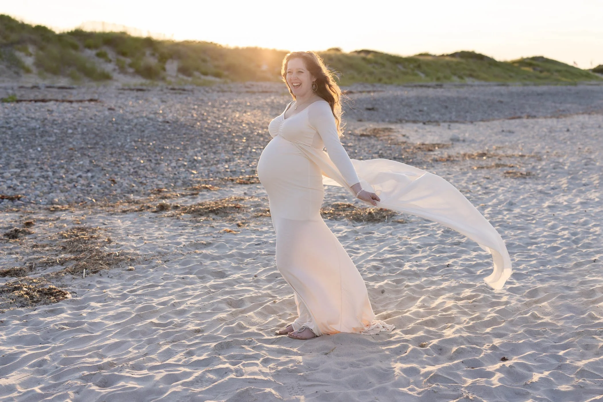 Happy pregnant woman standing on beach wearing cream colored dress flowing beautifully in breeze backlit by setting sun