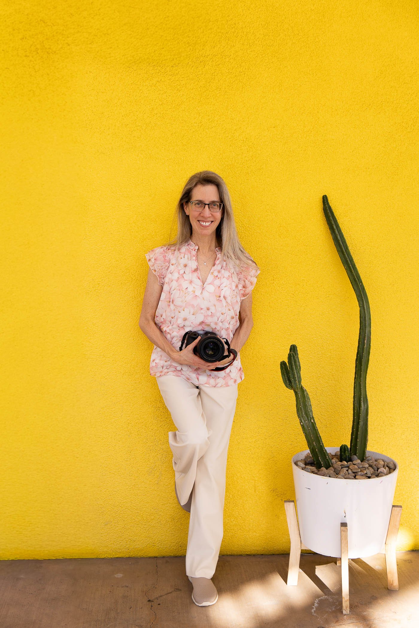 Portrait of Jennifer Banks smiling holding Canon camera wearing soft pink blouse and light tan pants.