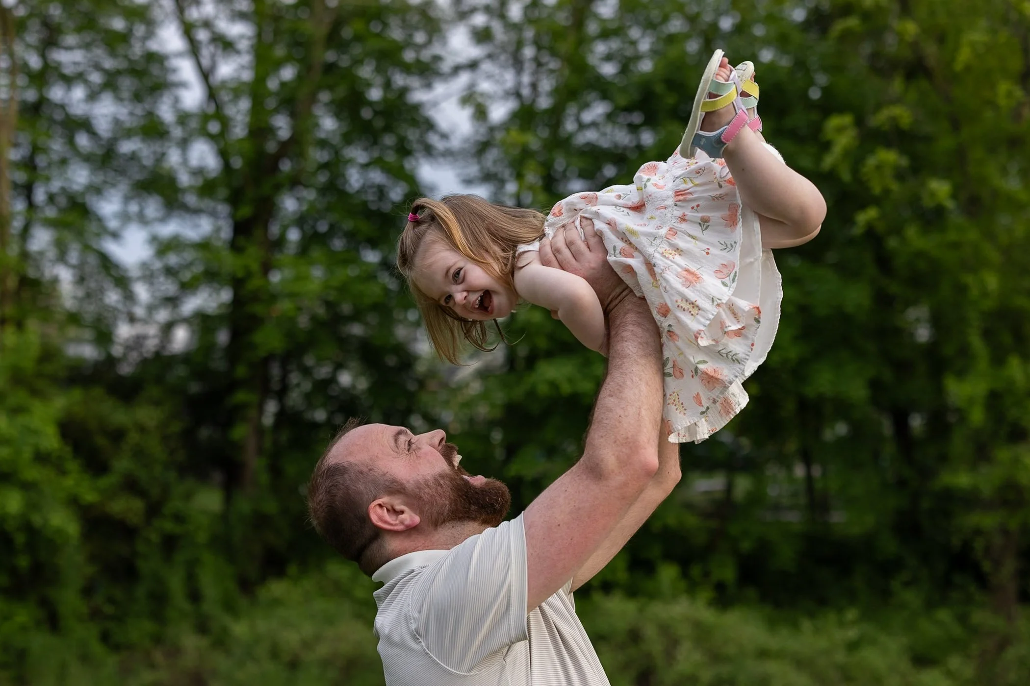 Dad with beard smiles and tosses young daughter outdoors in park little girl wearing white dress with peach colored flowers