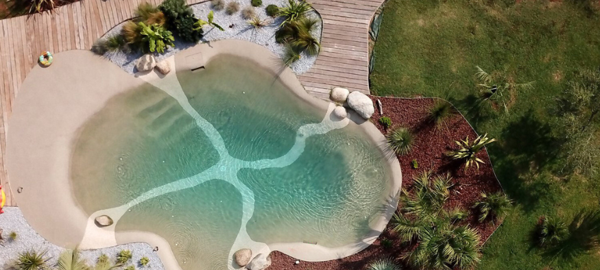 A backyard with a freeform swimming pool surrounded by a wooden deck, rocks, and desert plants, with grassy areas and trees nearby.