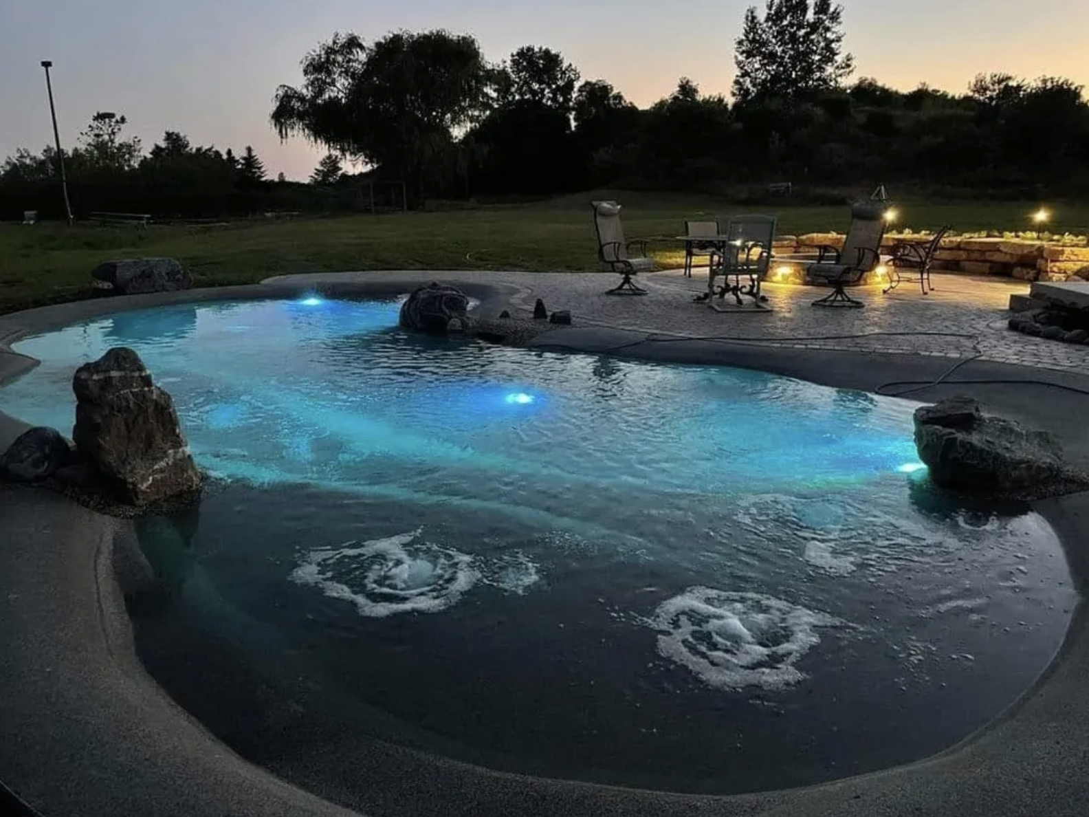 A backyard pool area at dusk with illuminated water, outdoor chairs, a table, a stone wall with lights, and trees in the background.