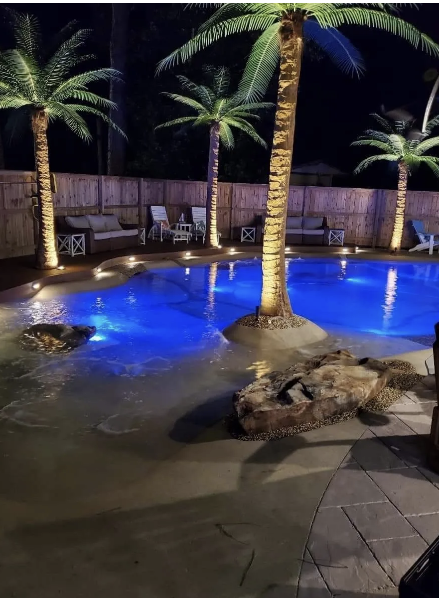 Nighttime backyard scene featuring a small illuminated swimming pool with palm trees and outdoor furniture along the wooden fence.