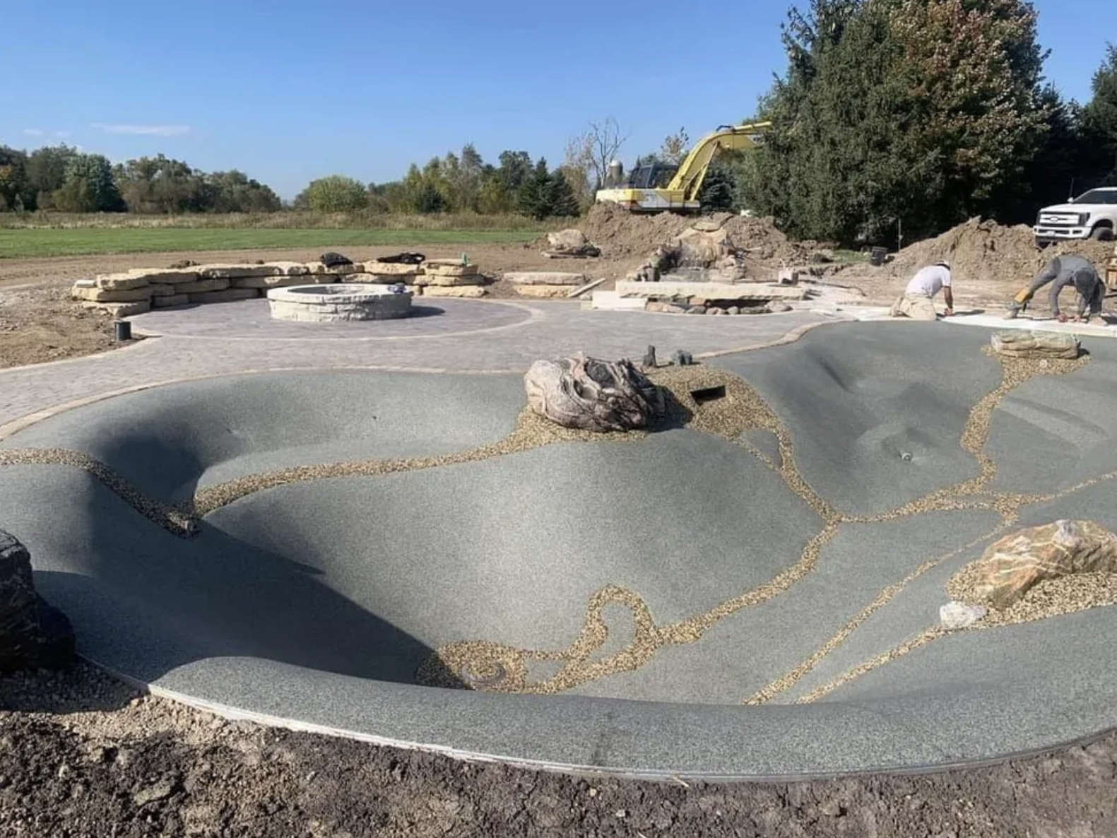 Under construction outdoor area with a small artificial pond, paved walkways, a fire pit, rocks, and a yellow excavator in the background.