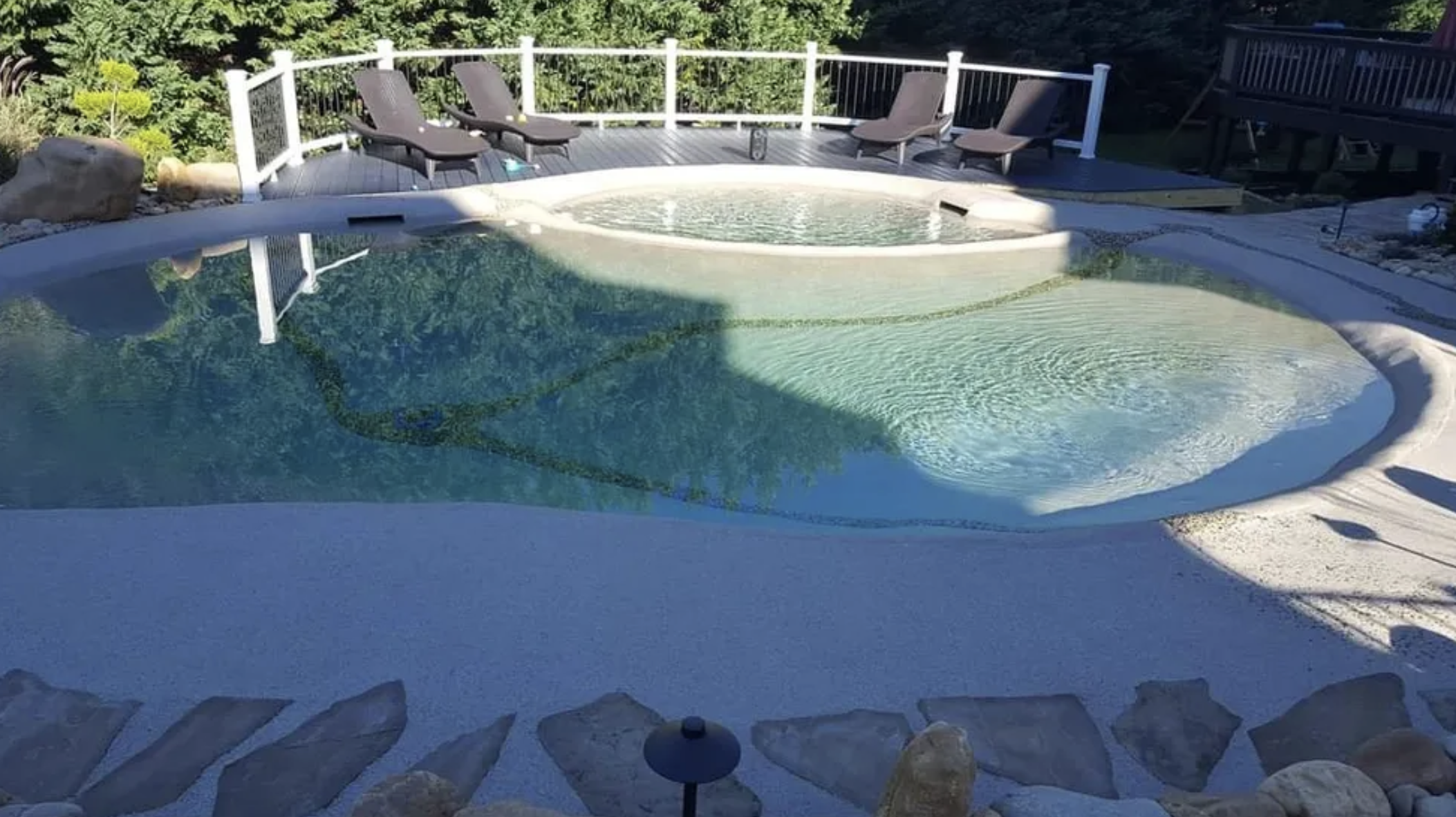 A backyard swimming pool with a circular hot tub attached, surrounded by a deck with lounge chairs and a white fence