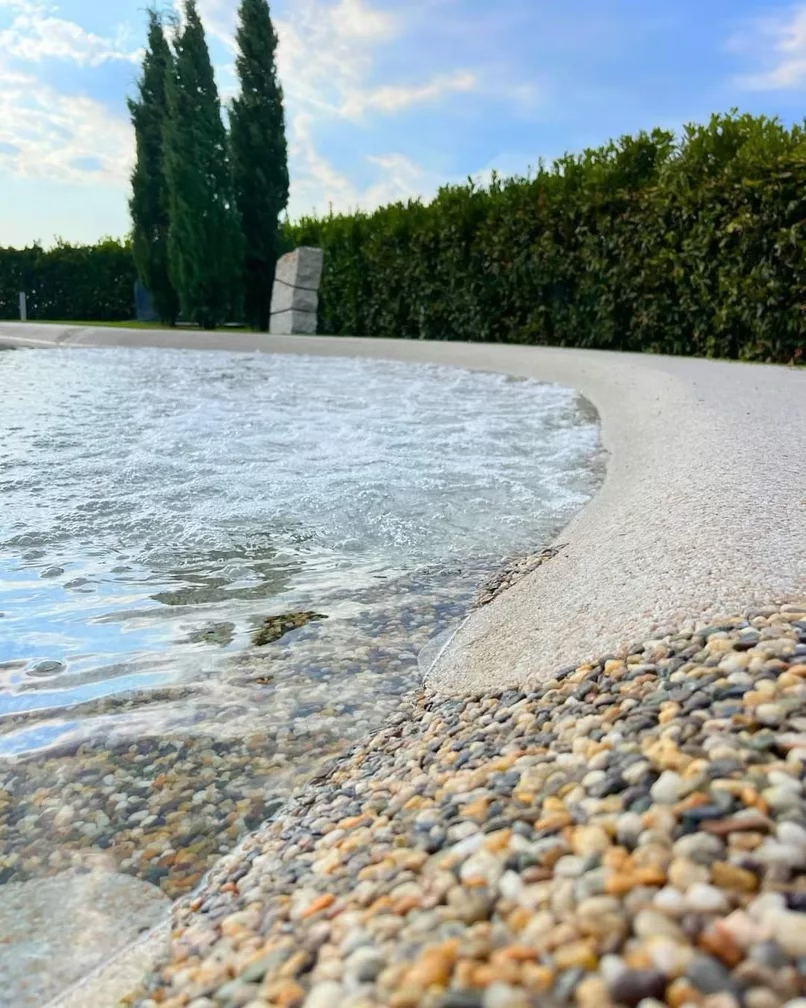 Close-up view of a beach with pebbles along the shoreline, water gently lapping onto the sand, with a hedge, tall trees, and a partly cloudy sky in the background.