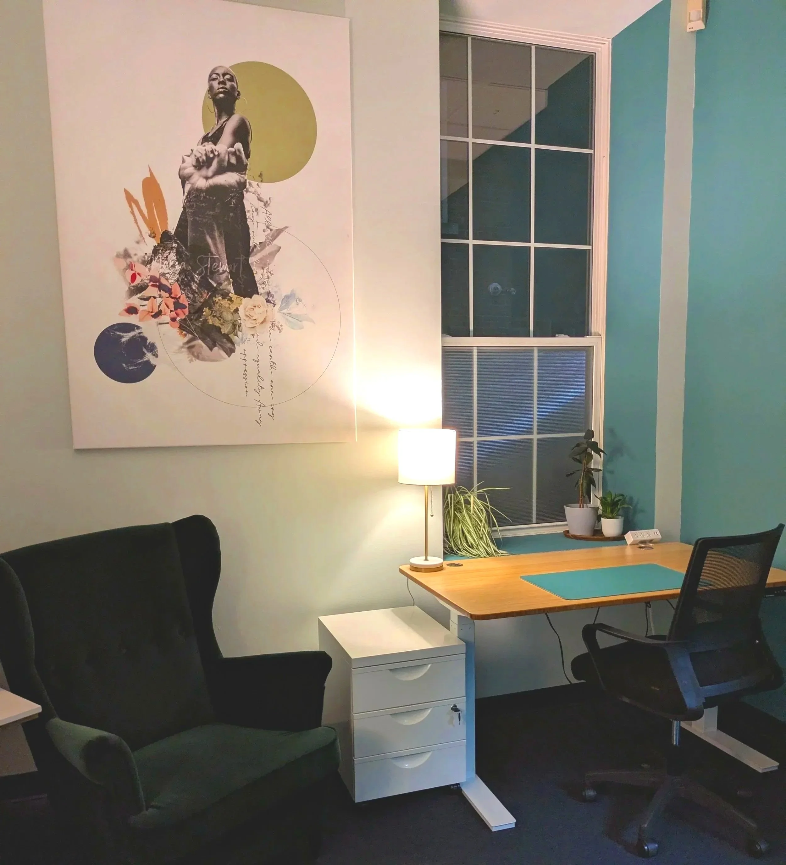 A cozy home office with a black upholstered armchair, a wooden desk with a blue desk mat, a white desk drawer unit, a small white shelf, a tall floor lamp, and a window with potted plants. A large artistic poster hangs on the wall. CT Coworking