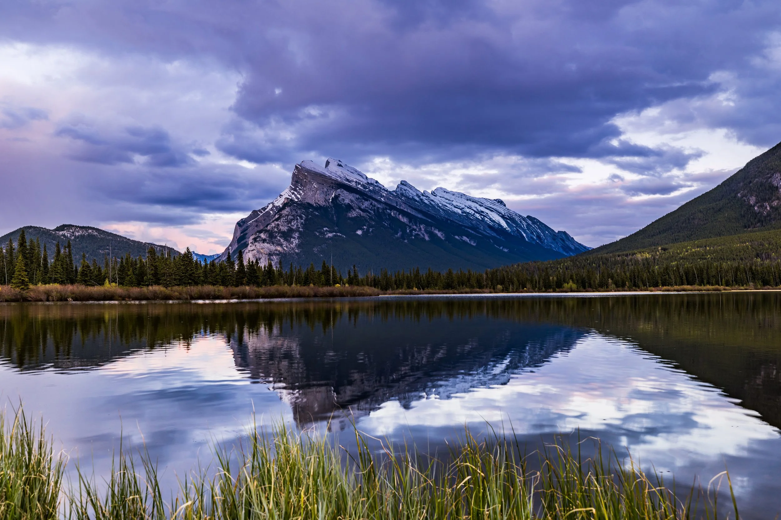 A mountain landscape showing a snow-capped mountain reflected in a calm lake, with a forested shoreline and a cloudy sky.
