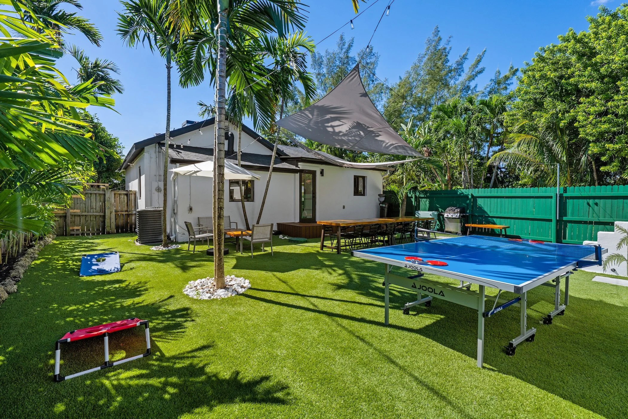 Backyard with a ping pong table, outdoor seating, a shade sail, and lush green trees and plants under a blue sky.