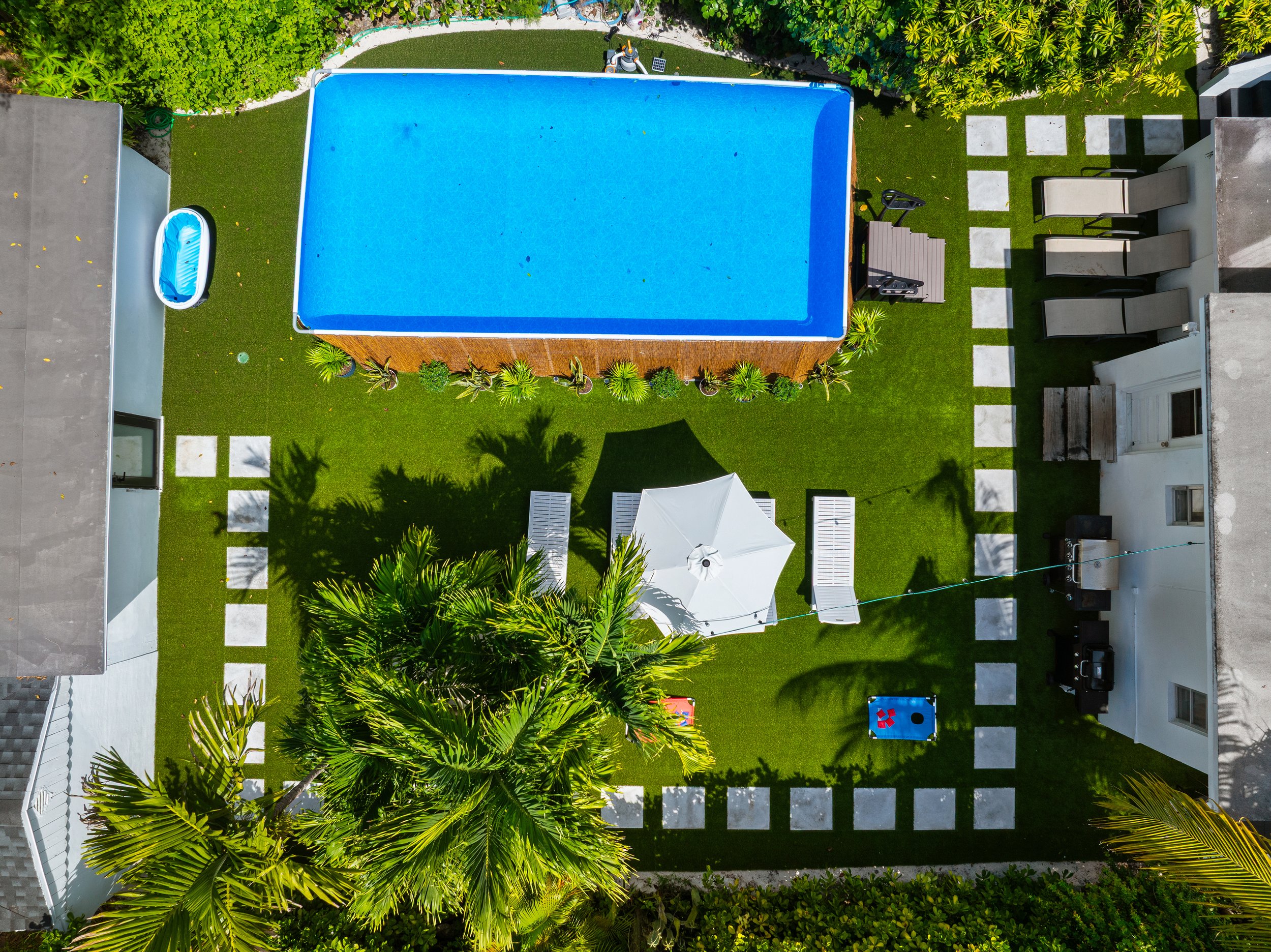 Aerial view of a backyard with a swimming pool, a small inflatable pool, a white umbrella shading two lounge chairs, a cornhole game, a ping pong table, and various potted plants.