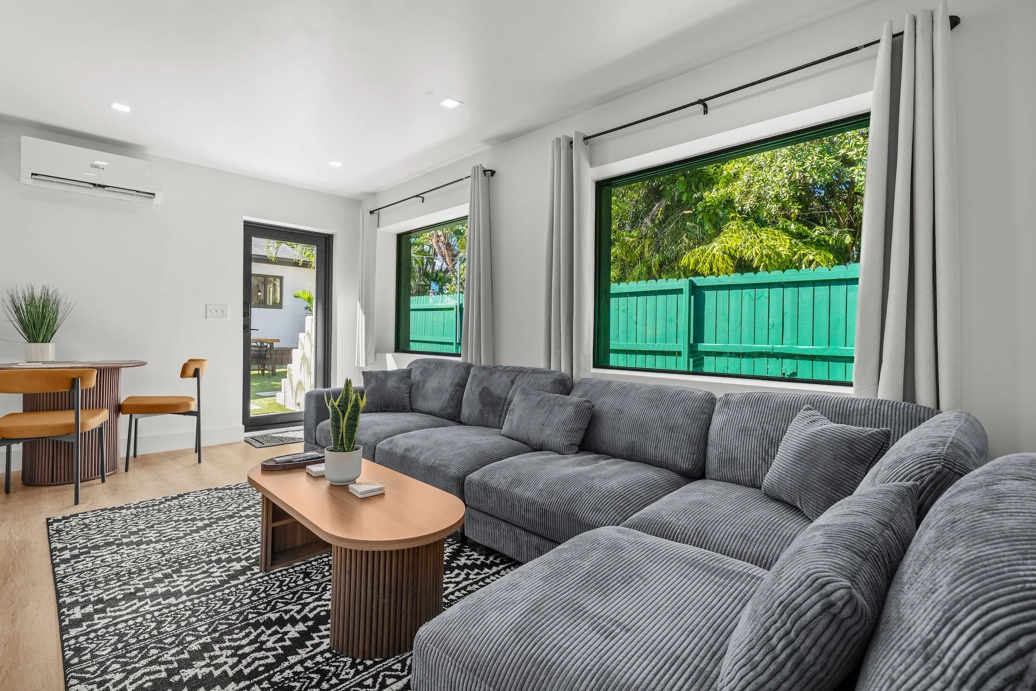 Living room with gray sectional sofa, wooden coffee table, black-and-white patterned rug, large windows with white curtains, and a sliding door leading to a backyard with green fencing and trees.