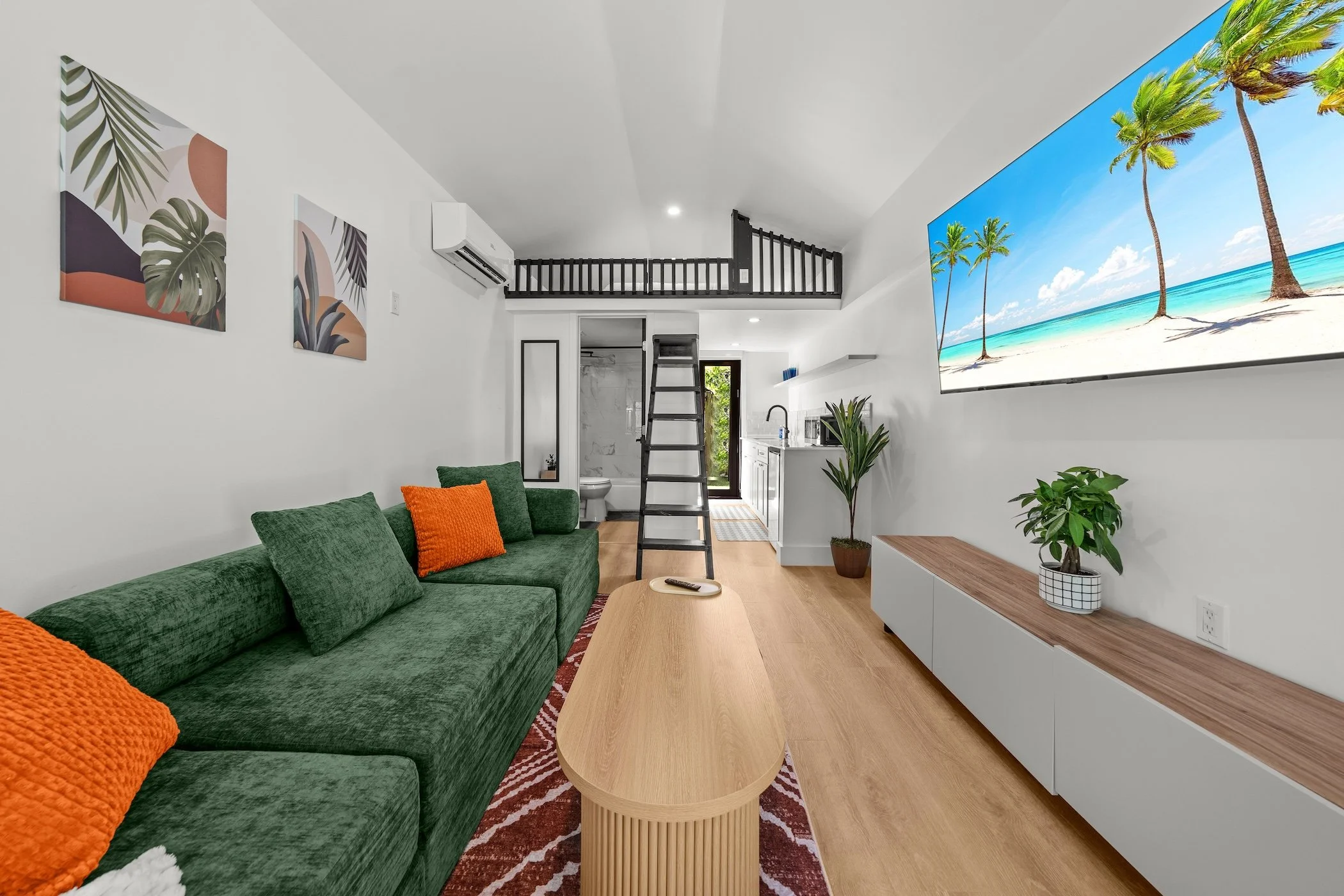 Modern living room with green couch, orange pillows, wooden coffee table, large wall-mounted TV showing beach scene, plants, and art on white walls