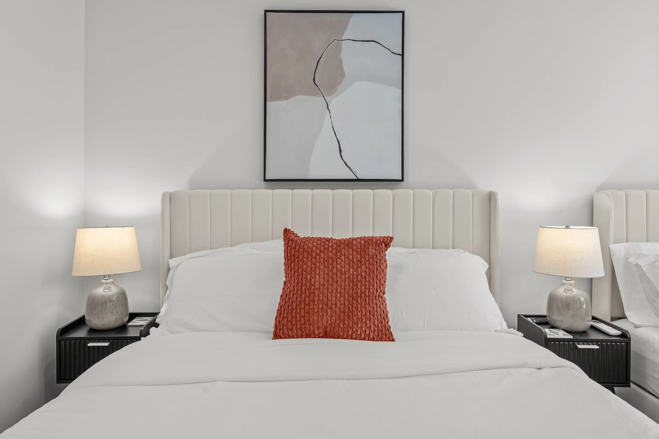 A neatly made bed with a white headboard, white bedding, and a red textured throw pillow in the center. There are two matching nightstands with table lamps on either side of the bed. A piece of abstract art hangs above the headboard.