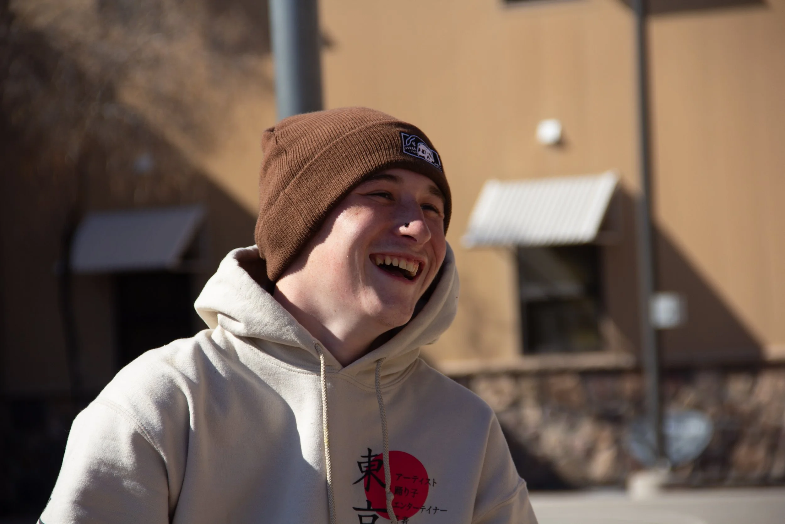 A young man wearing a brown beanie and a cream hoodie, smiling and laughing outdoors on a sunny day.