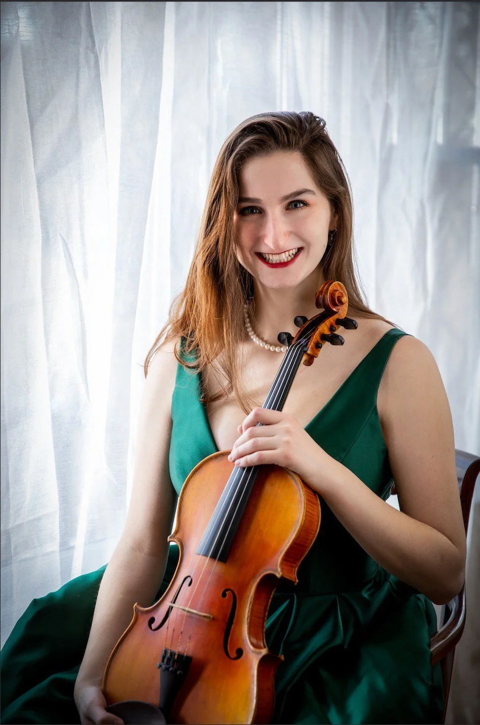 A young woman with long brown hair, wearing a green dress and pearl necklace, sitting and smiling while holding a violin.