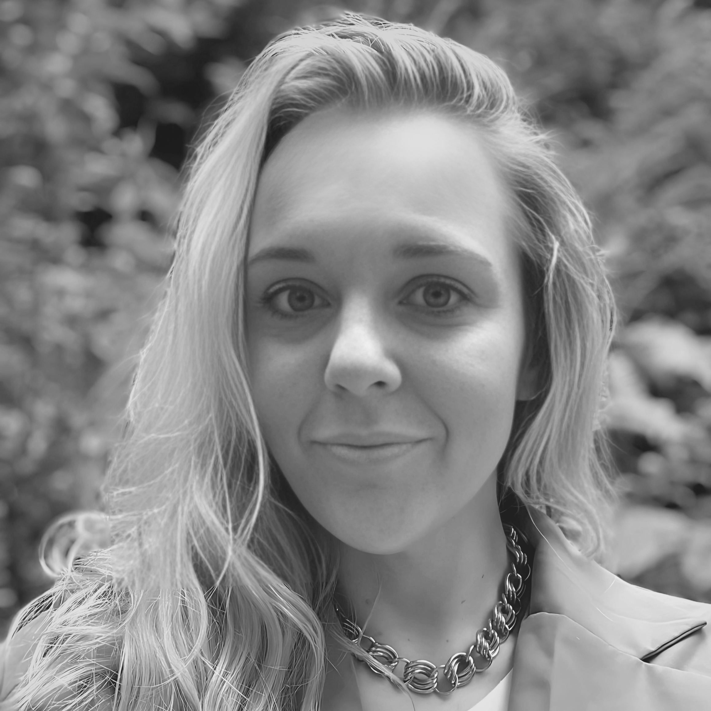 A woman with long wavy hair, wearing a chain necklace and a jacket, smiling softly, outdoors with blurred natural background.