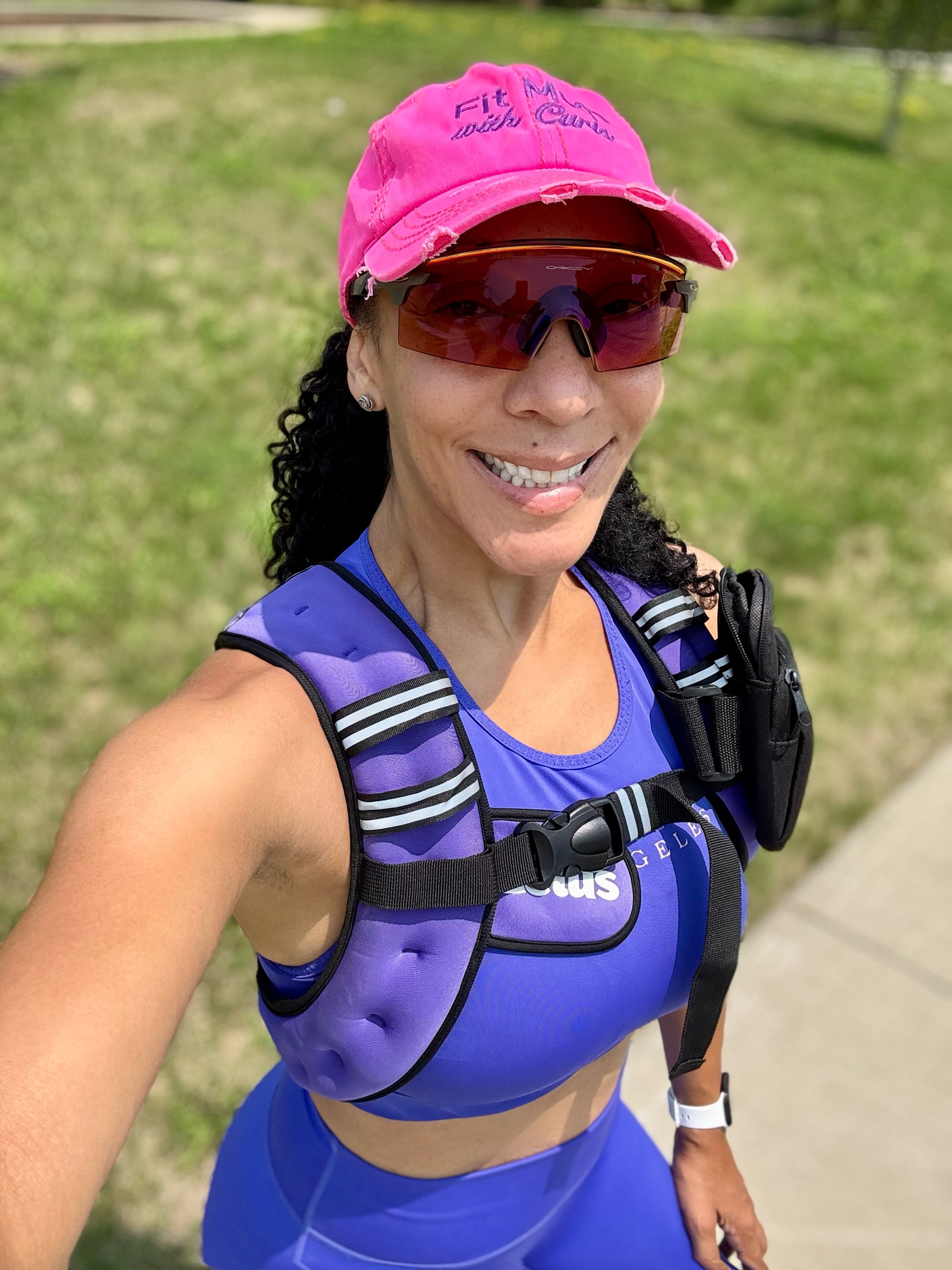 A woman in athletic attire, wearing a pink cap, sunglasses, and a purple sports bra, taking a selfie outdoors on a sunny day.