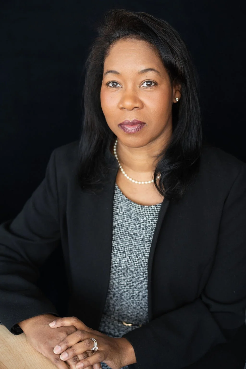 A professional woman with dark hair and a serious expression, wearing a black blazer, pearl necklace, and earrings, seated at a desk with a black background.