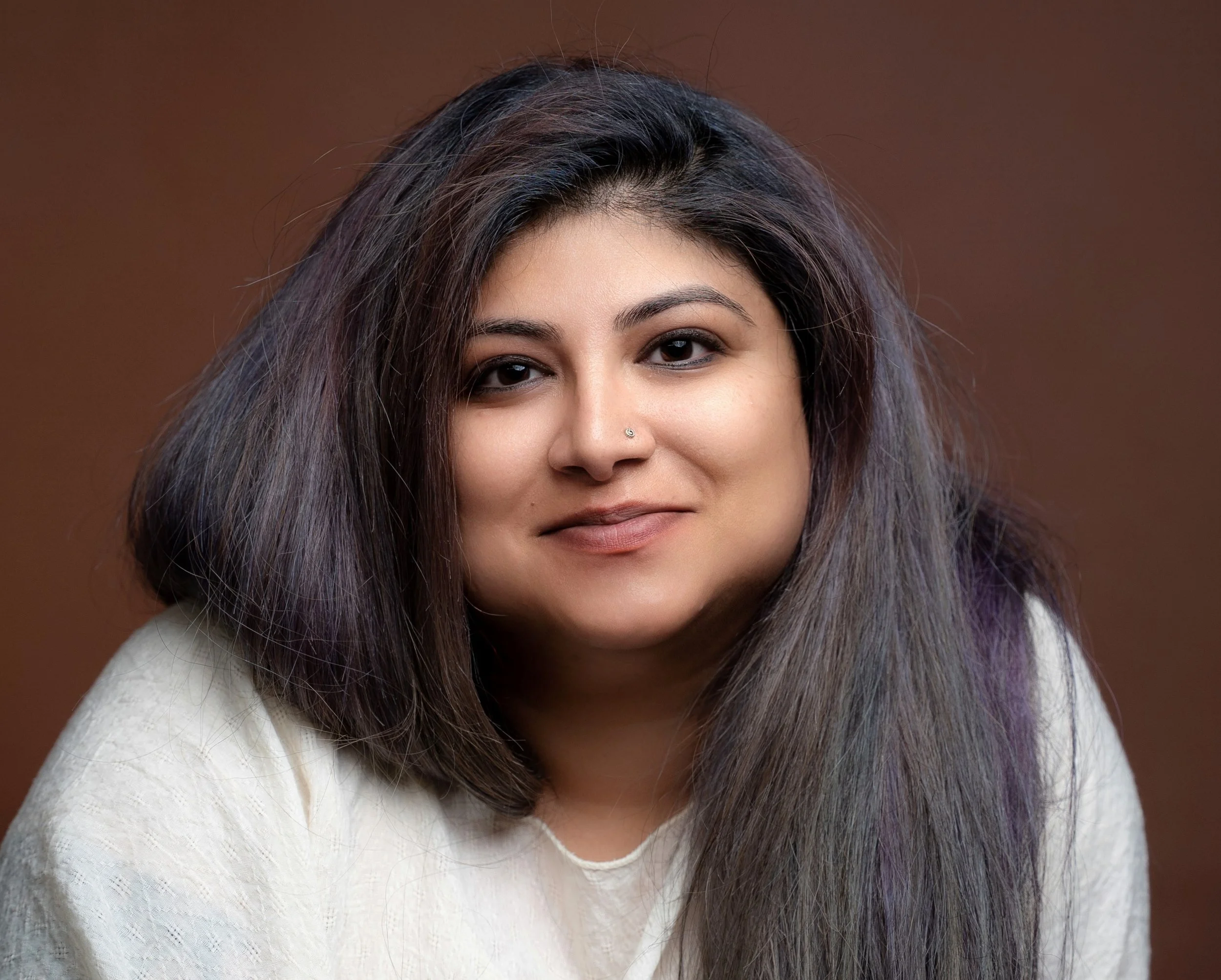A woman with long, dark, tousled hair and light skin, smiling softly, wearing a white top, with a brown neutral background.