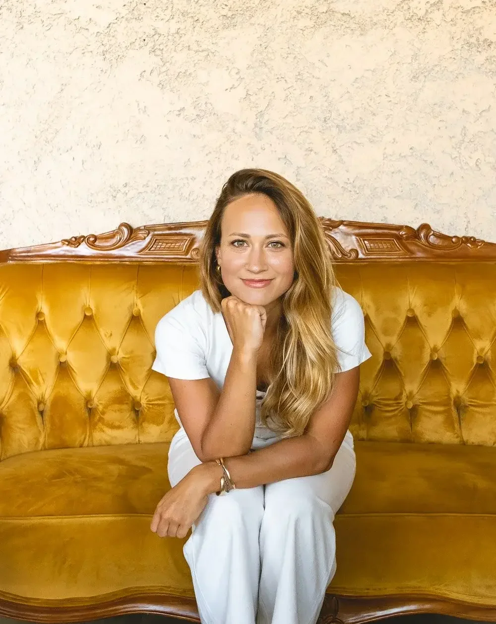 Katy sitting on a vintage gold tufted sofa with wooden detailing, in front of a textured beige wall.
