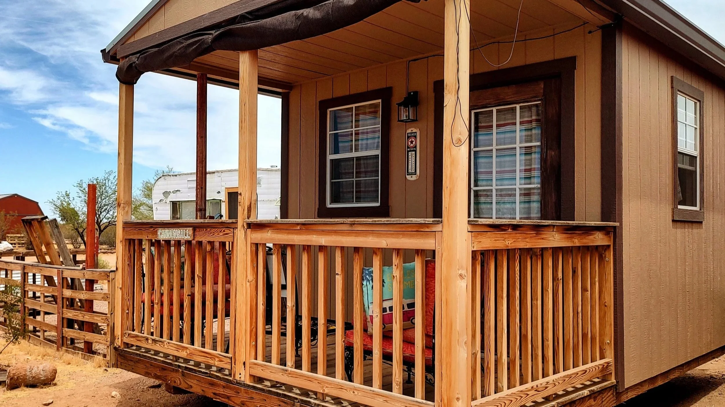 exterior image of a cabin structure showing a wooden railing a window and door with striped curtains. a wooden fence is seen on the left. blue sky with some clouds.