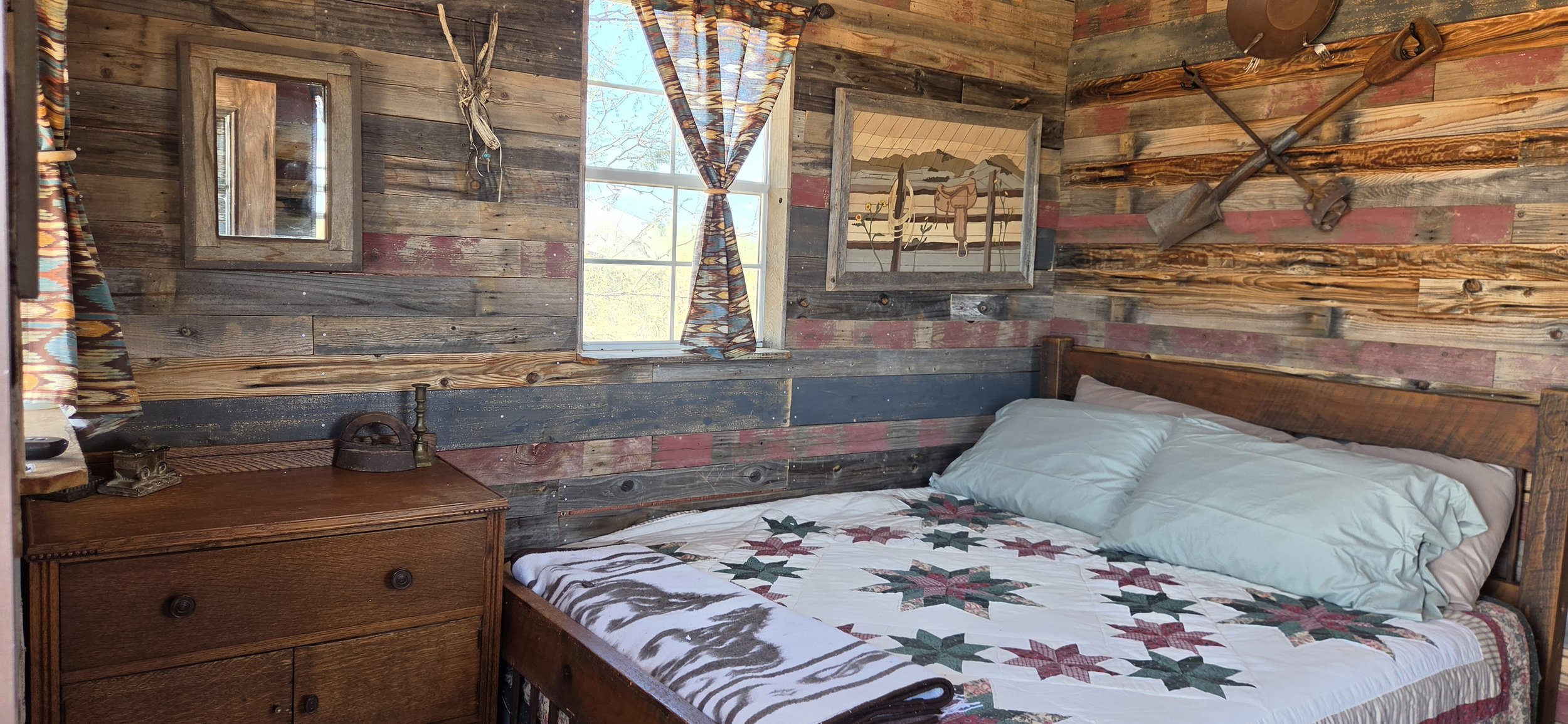 Rustic bedroom with wood panel walls, a window with plaid curtains, a wall-mounted mirror, and vintage tools hanging on the wall. The bed has a quilt with holiday patterns and a pillow, next to a wooden nightstand with an old iron and a small decorative item.