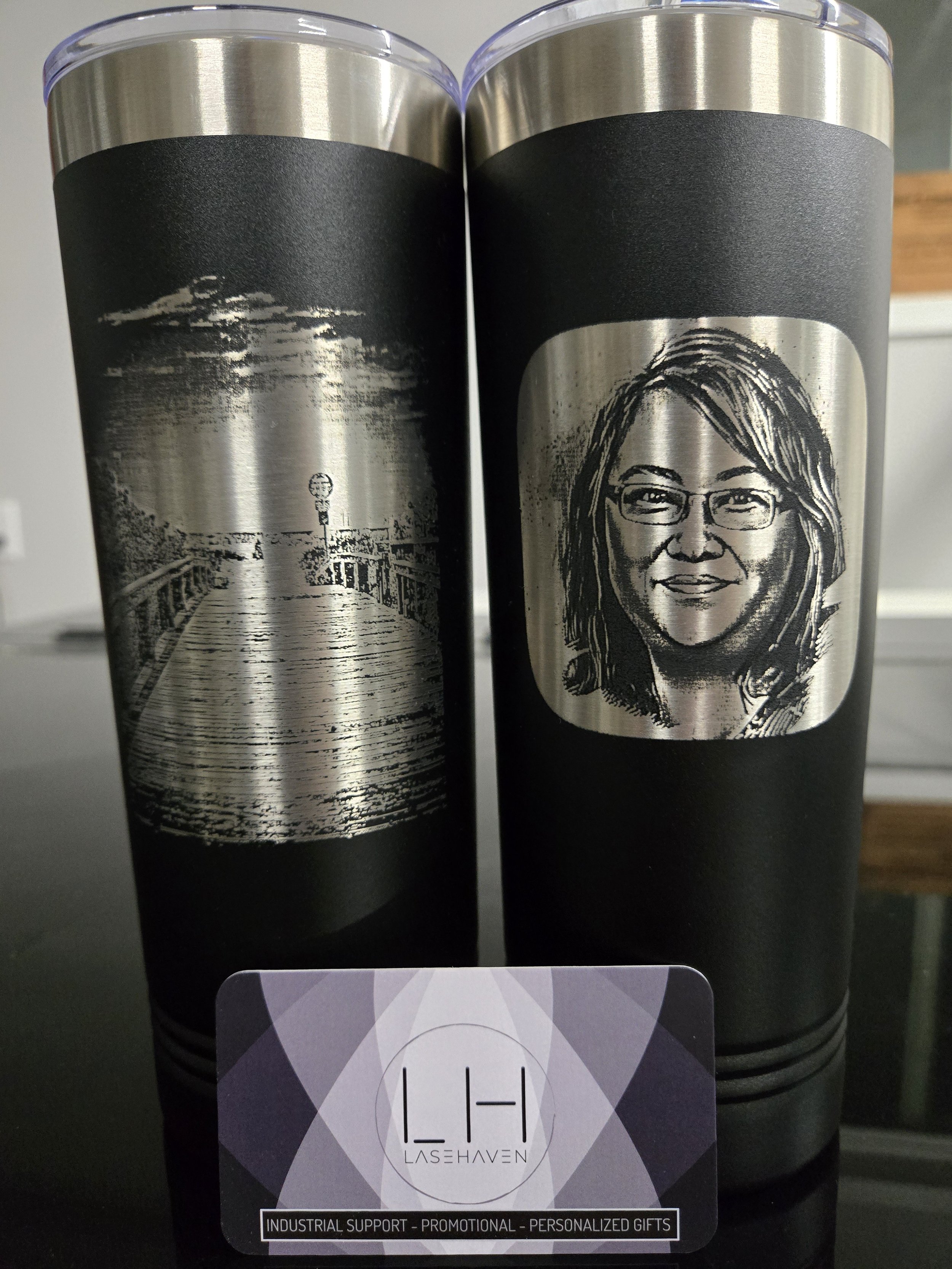 Two black and stainless steel tumblers with laser etched designs; one with a landscape scene, the other with a portrait of a person. In front, a business card with "LaseHaven" logo and text: "Industrial Support - Promotional - Personalized Gifts."