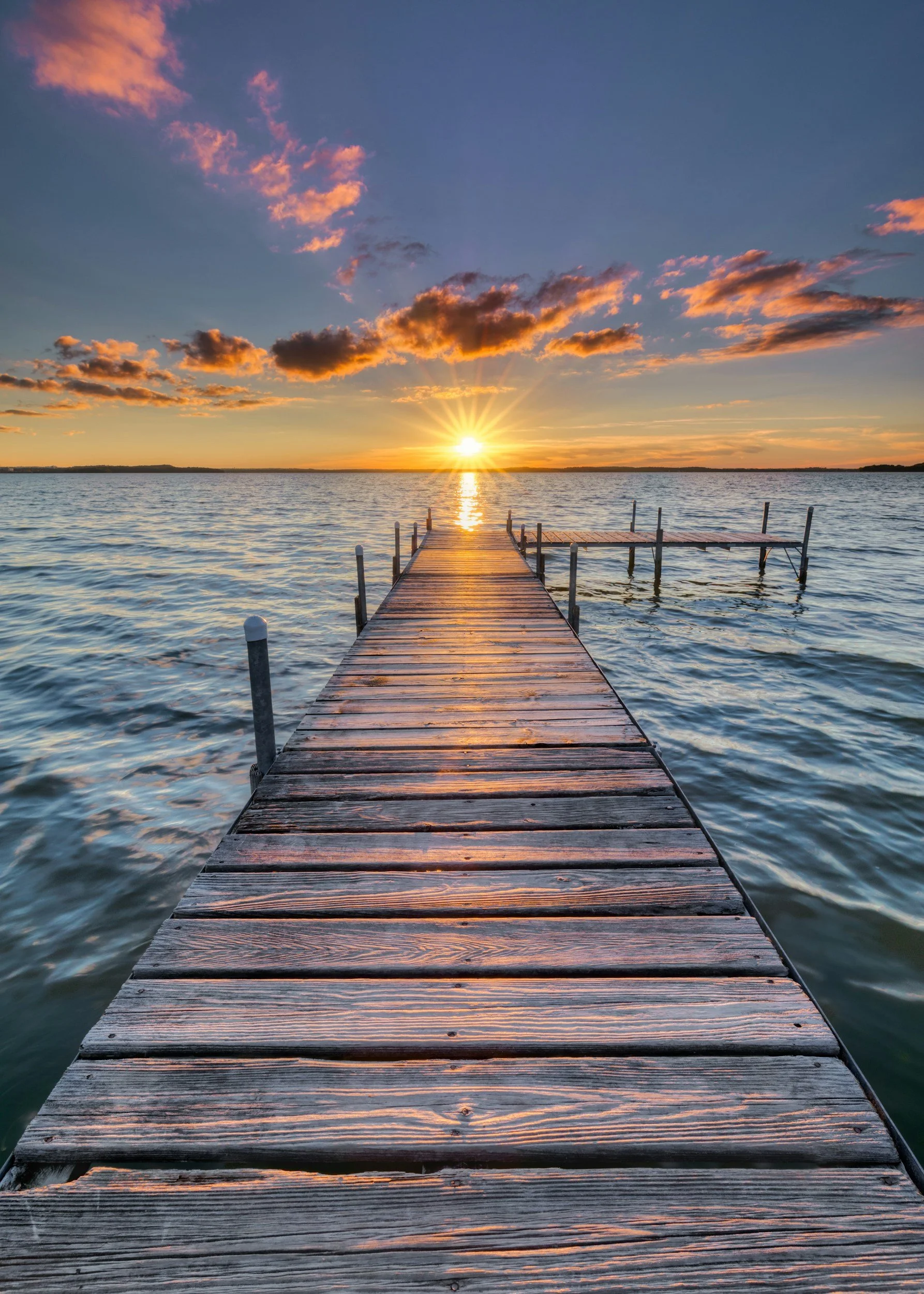 A wooden dock extending into a large body of water at sunset, with the sun near the horizon casting a warm glow, and clouds in the sky.