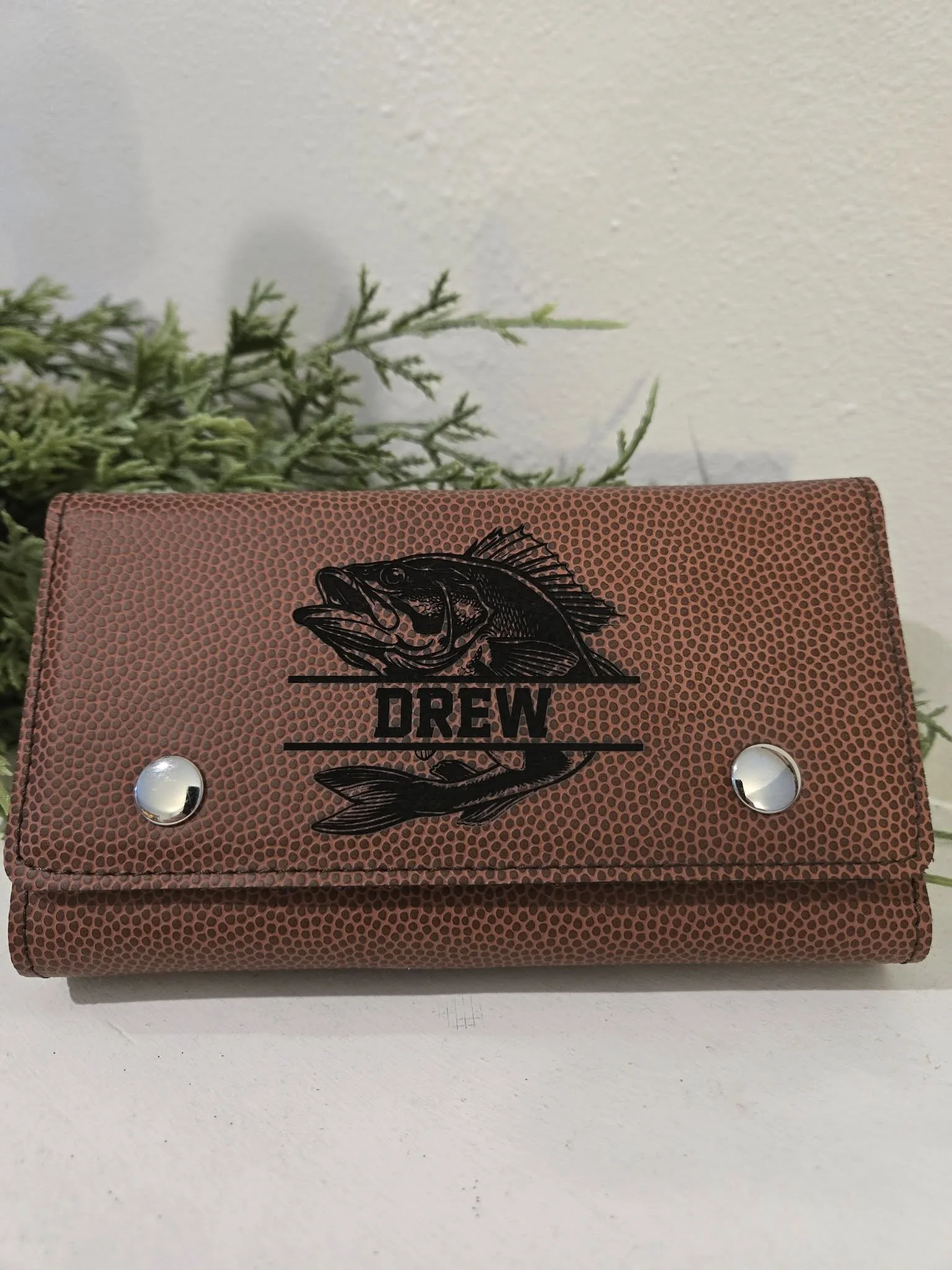 Brown leather wallet with fish logo and name 'Drew' embossed. Two metal snap closures, placed on a white surface with greenery in the background.