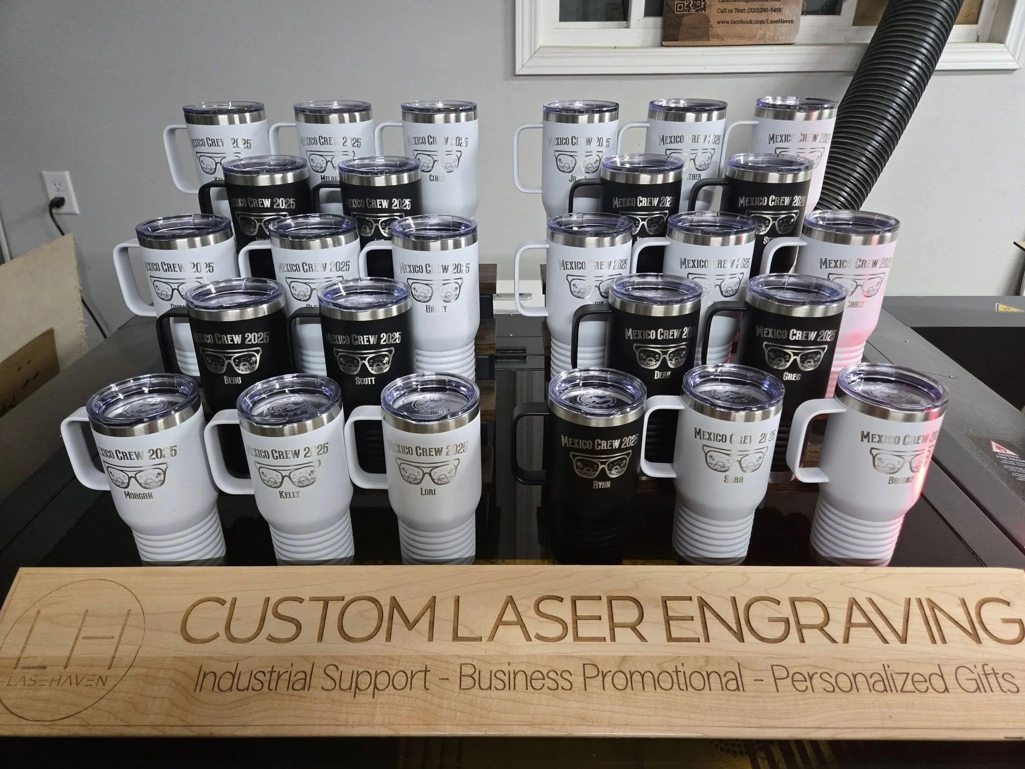 Several custom laser engraved travel mugs in black and white, labeled 'Mexico Crew 2025' with names, displayed on a table. A wooden sign reads 'Custom Laser Engraving' with details about industrial support, business promotional, and personalized gift
