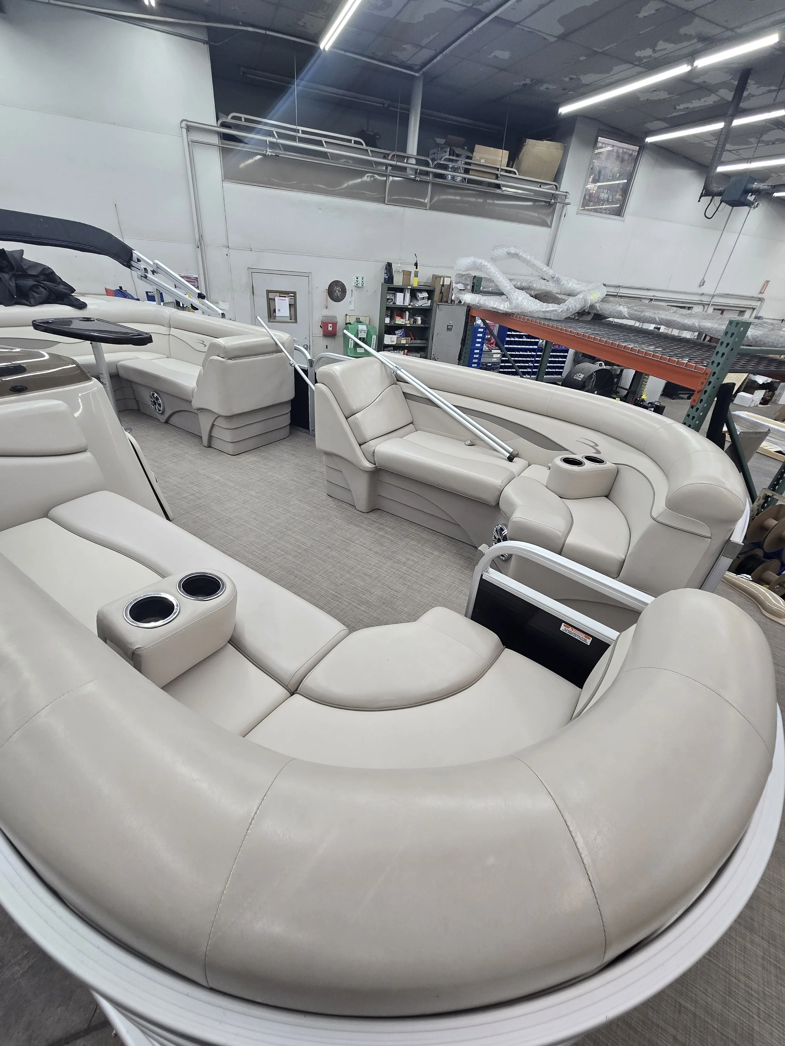A beige pontoon boat with multiple cushioned seating areas, cup holders, and a vinyl deck in a storage shed.