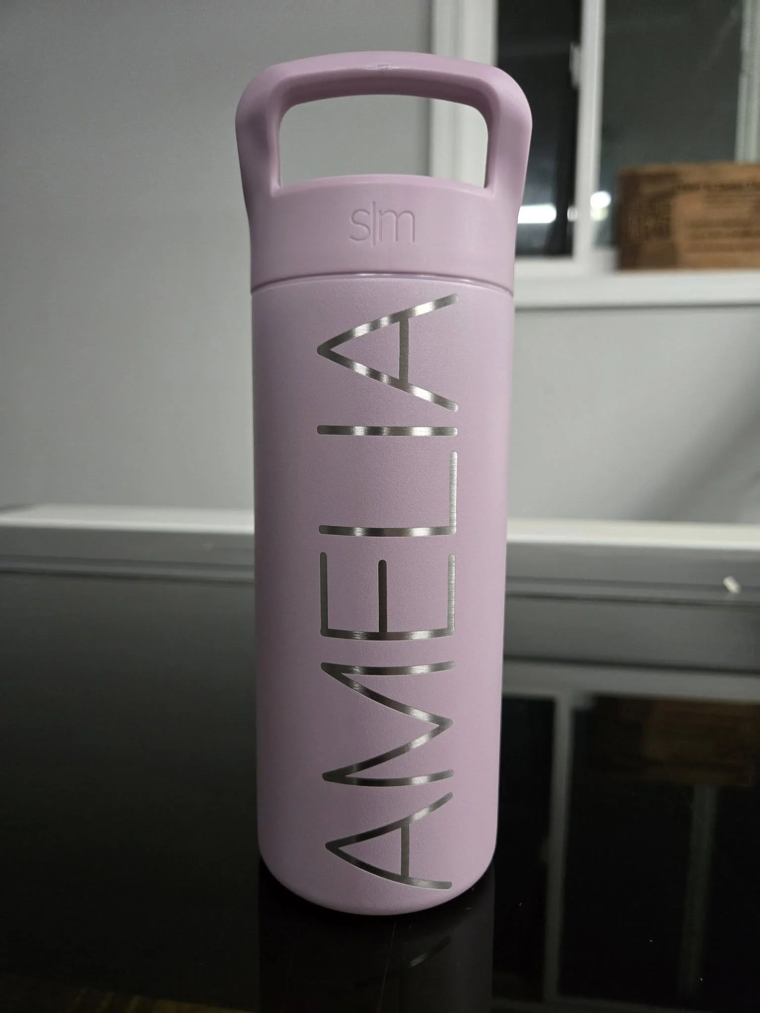 Purple water bottle with the word 'AMELIA' in metallic letters on a reflective surface, window in the background.