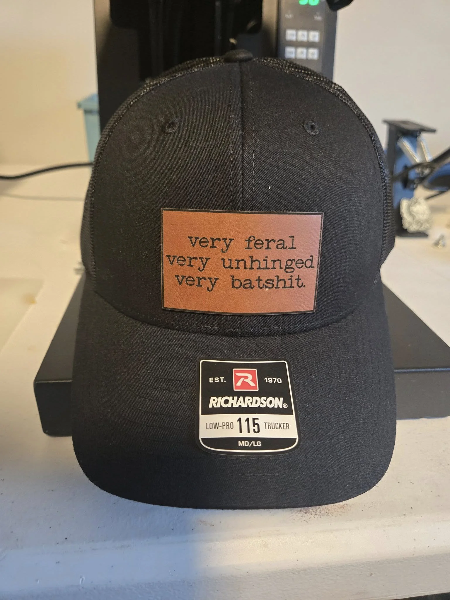 Black Richardson 115 Trucker hat with patch reading 'very feral, very unhinged, very batshit' in a room setting.