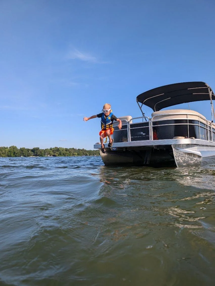 Child wearing goggles and life jacket jumping off paddle boat into lake on a sunny day.